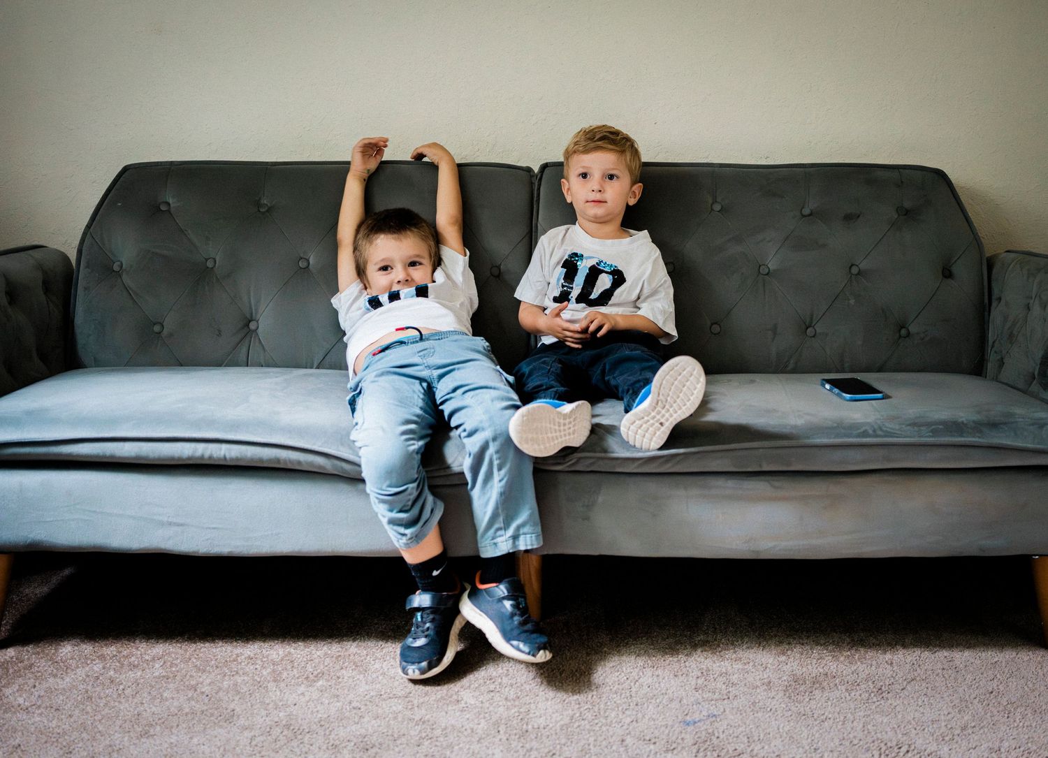 Two young children sit casually on a gray tufted couch in a living room.