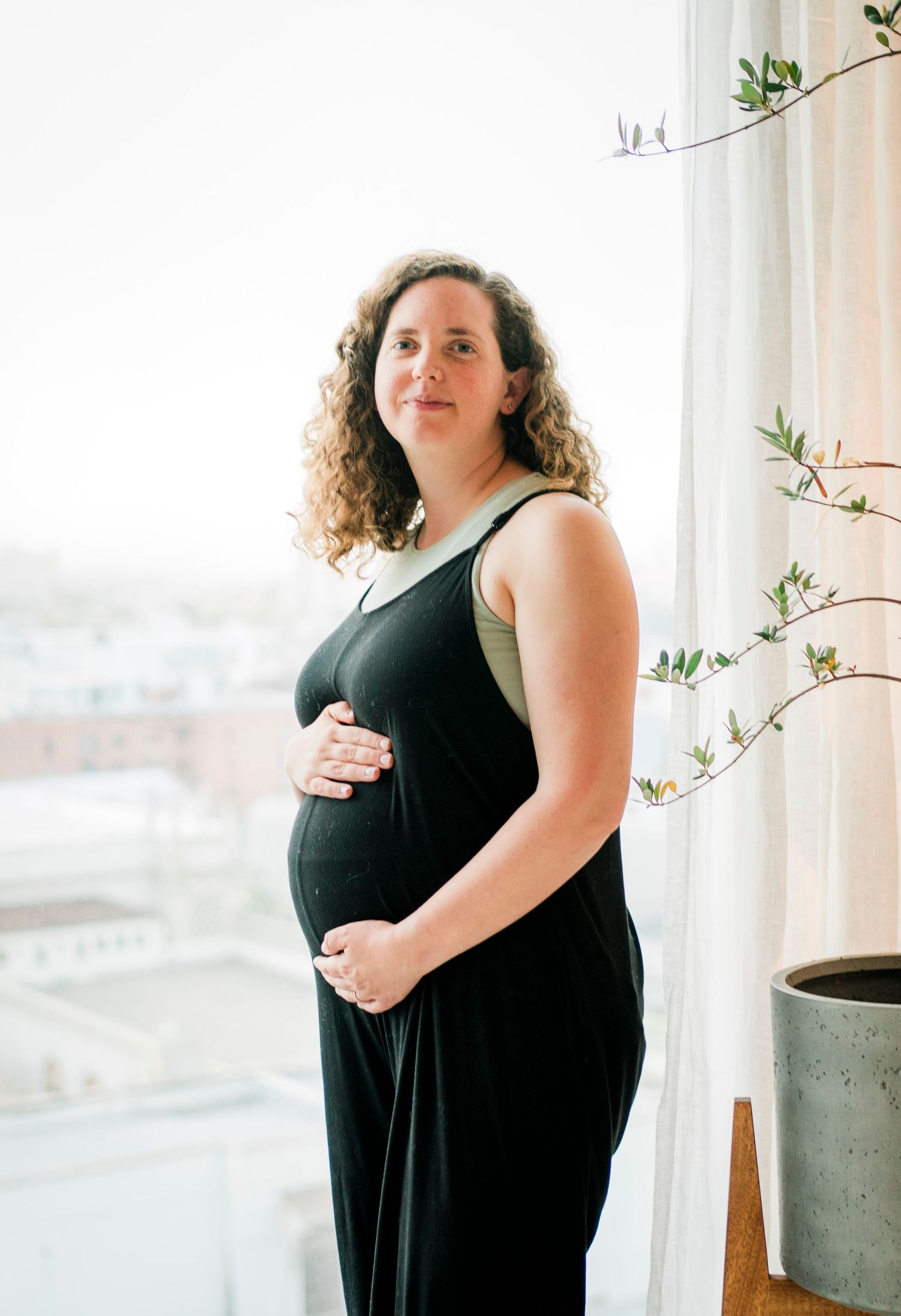 A maternity photo shoot featuring a black dress against a bright window with delicate plants.