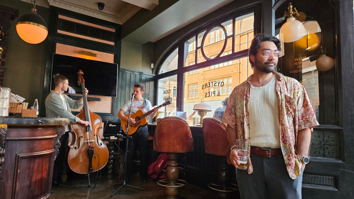 Warm interior of a rustic British pub with wooden beams and pendant lighting with a band in the background and man posed in the foreground.