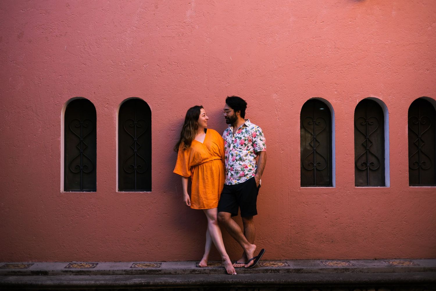 People walking past coral-colored wall with arched windows creating dramatic shadows.