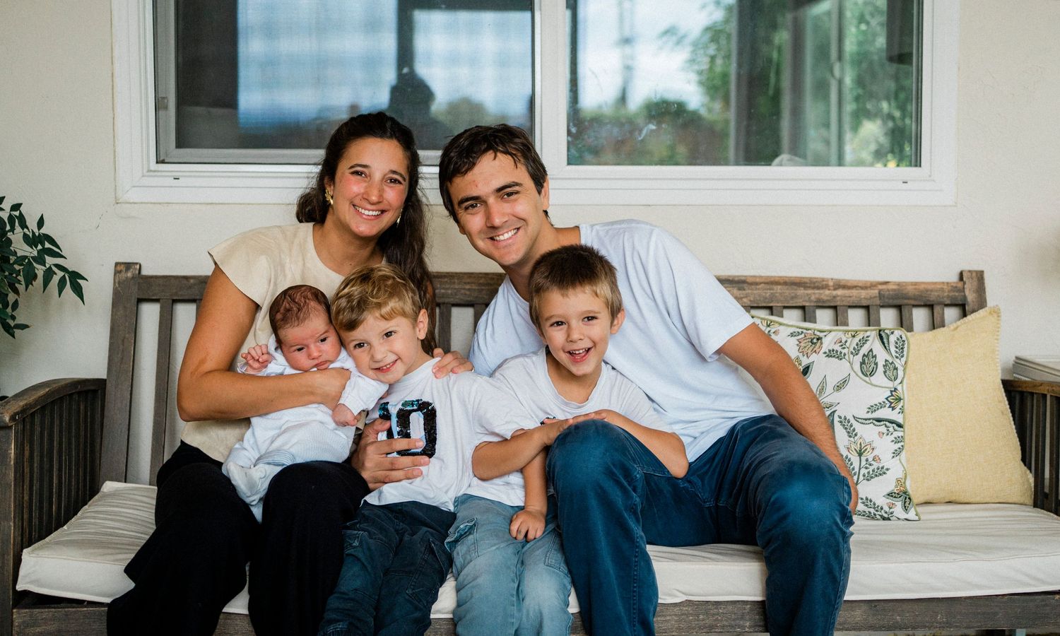 A happy family sits together on a white couch in their living room for a casual photo session.