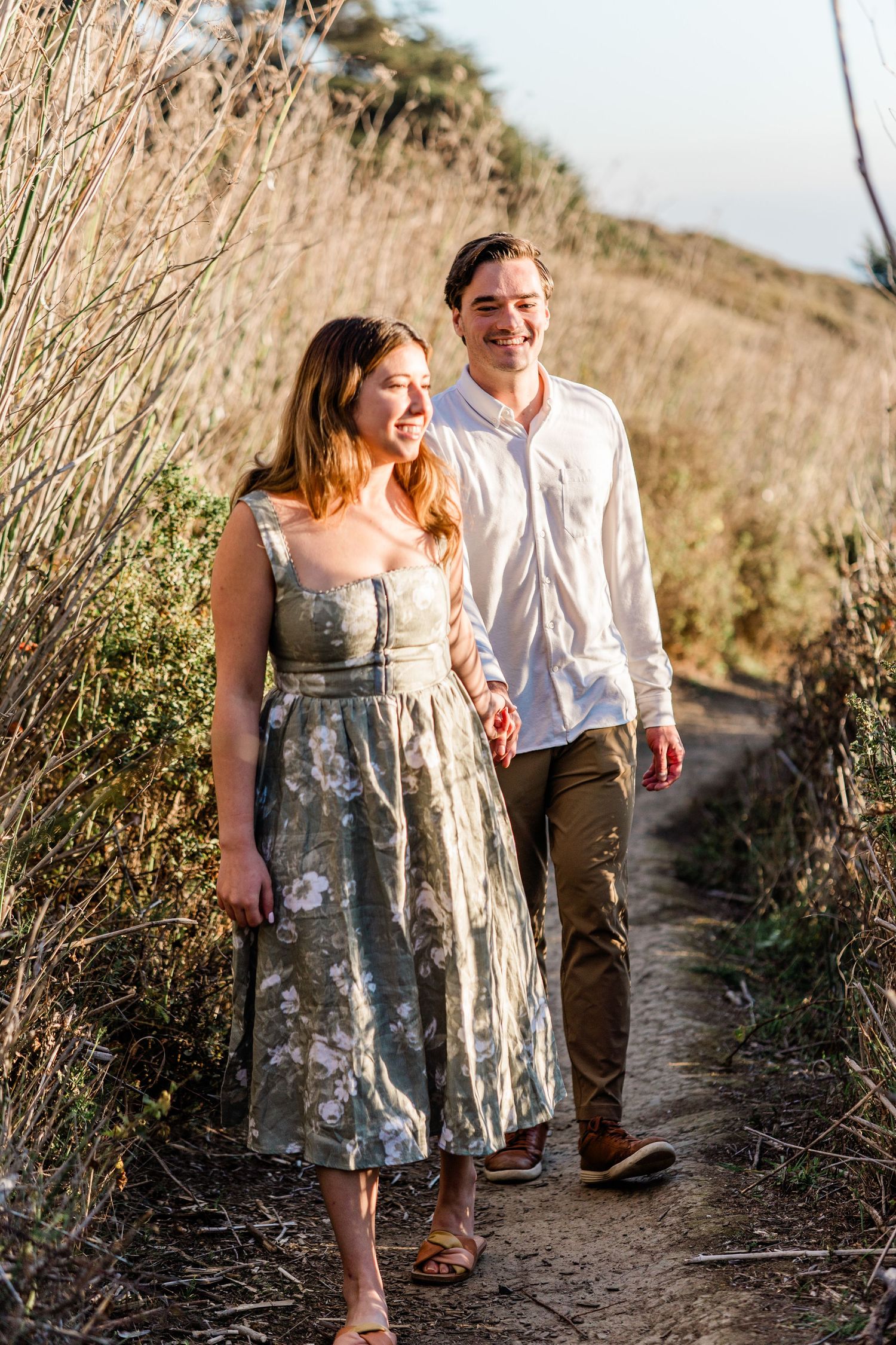 A couple walking hand in hand along a winding dirt path through dry grass on a hillside.