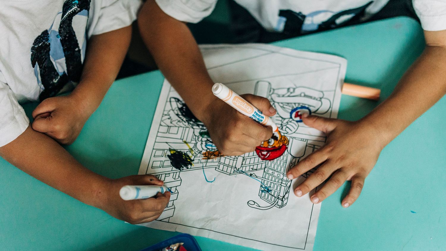 Close-up view of hands working on activity sheets or coloring pages on a turquoise surface.