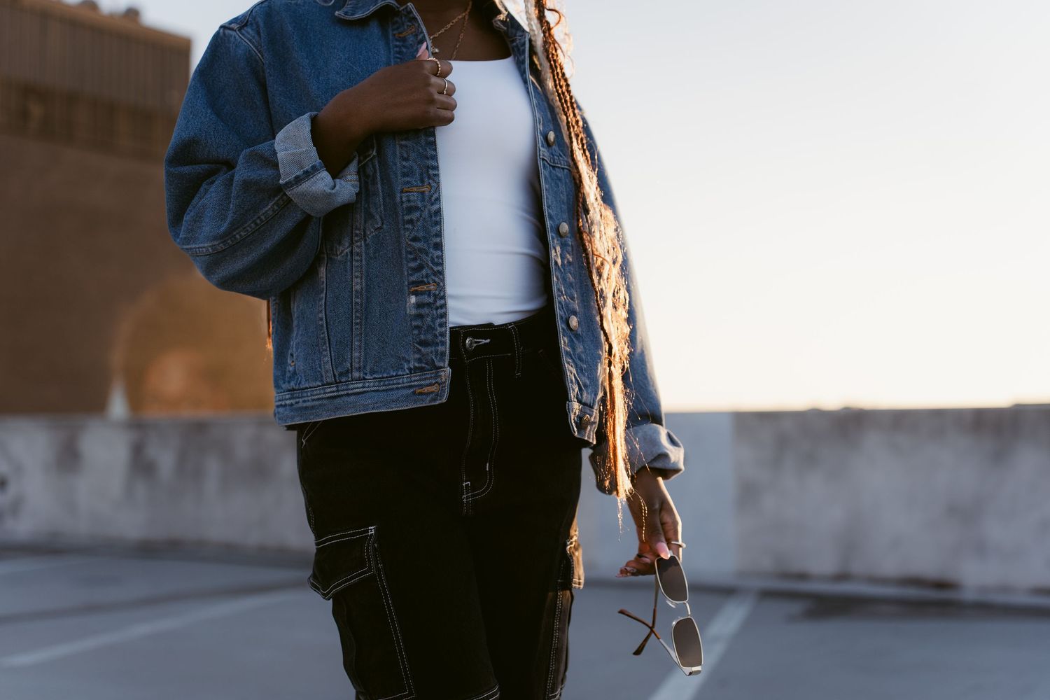 Portrait image of a woman’s body in a jean jacket, black pants, and holding sunglasses