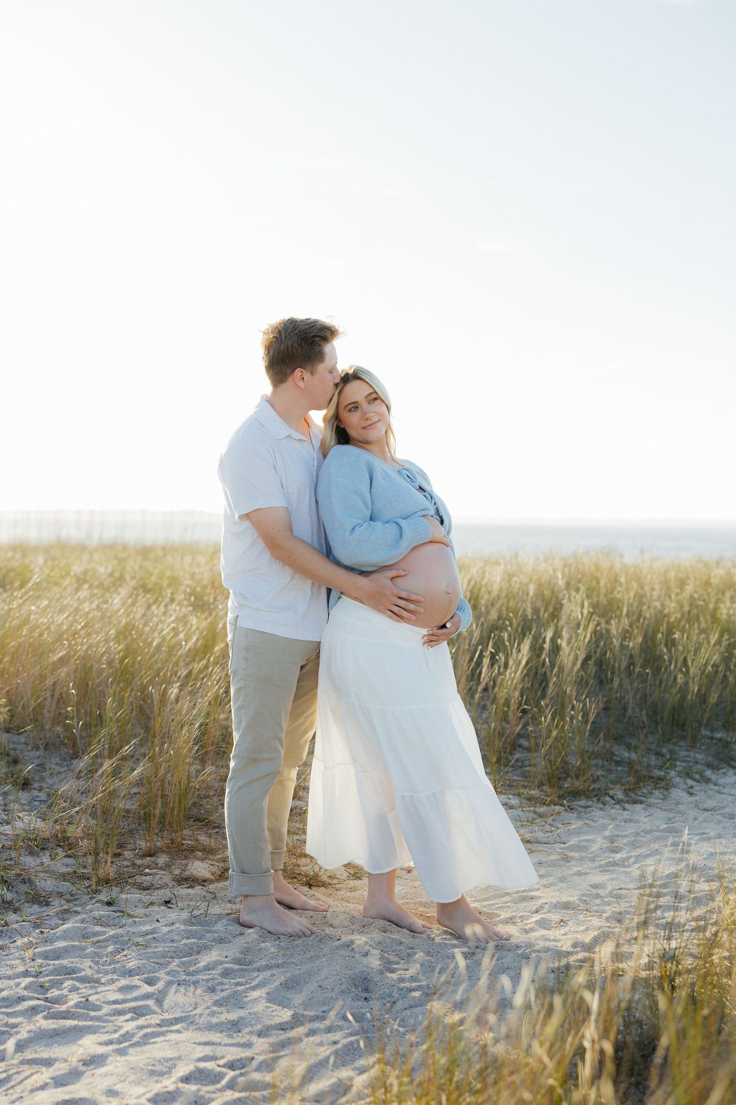 A romantic couple embraces on a sandy beach with sea grass and golden sunlight creating a dreamy coastal scene.