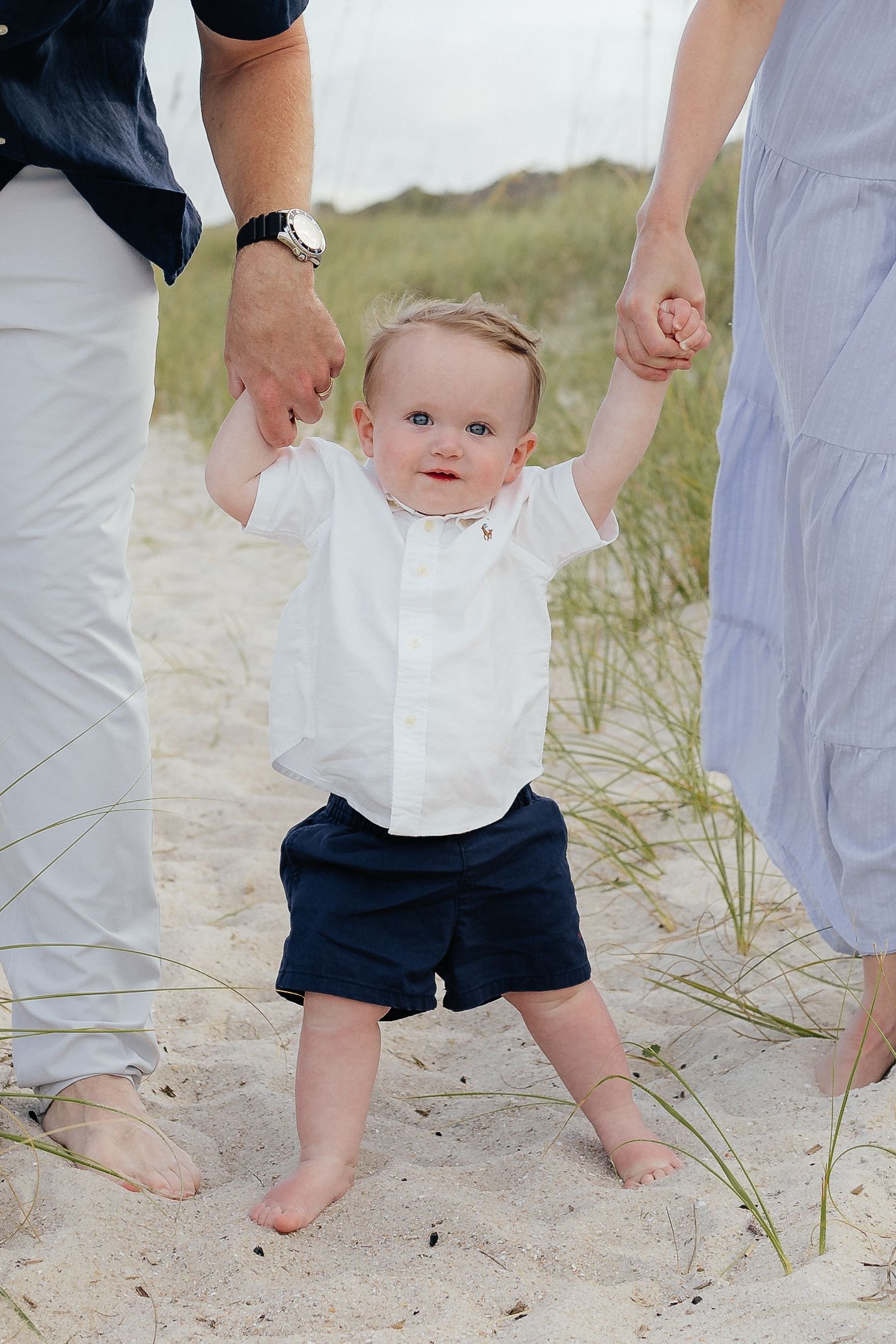 Small child in white shirt and navy shorts taking first steps on beach while holding parents' hands.