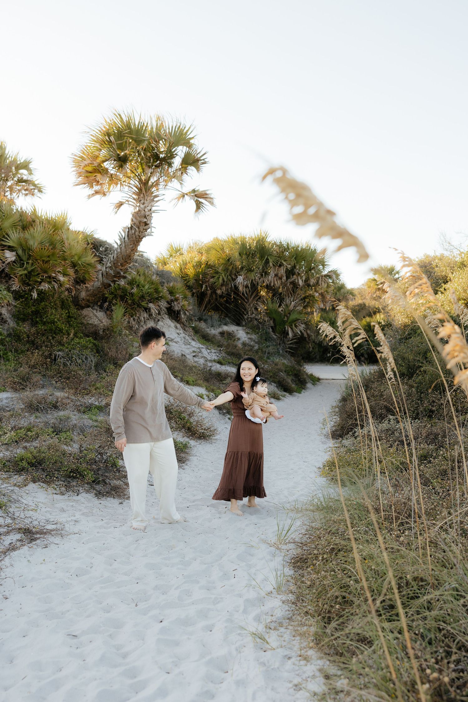 A couple walks along a sandy beach path surrounded by sea oats and coastal vegetation at sunset.