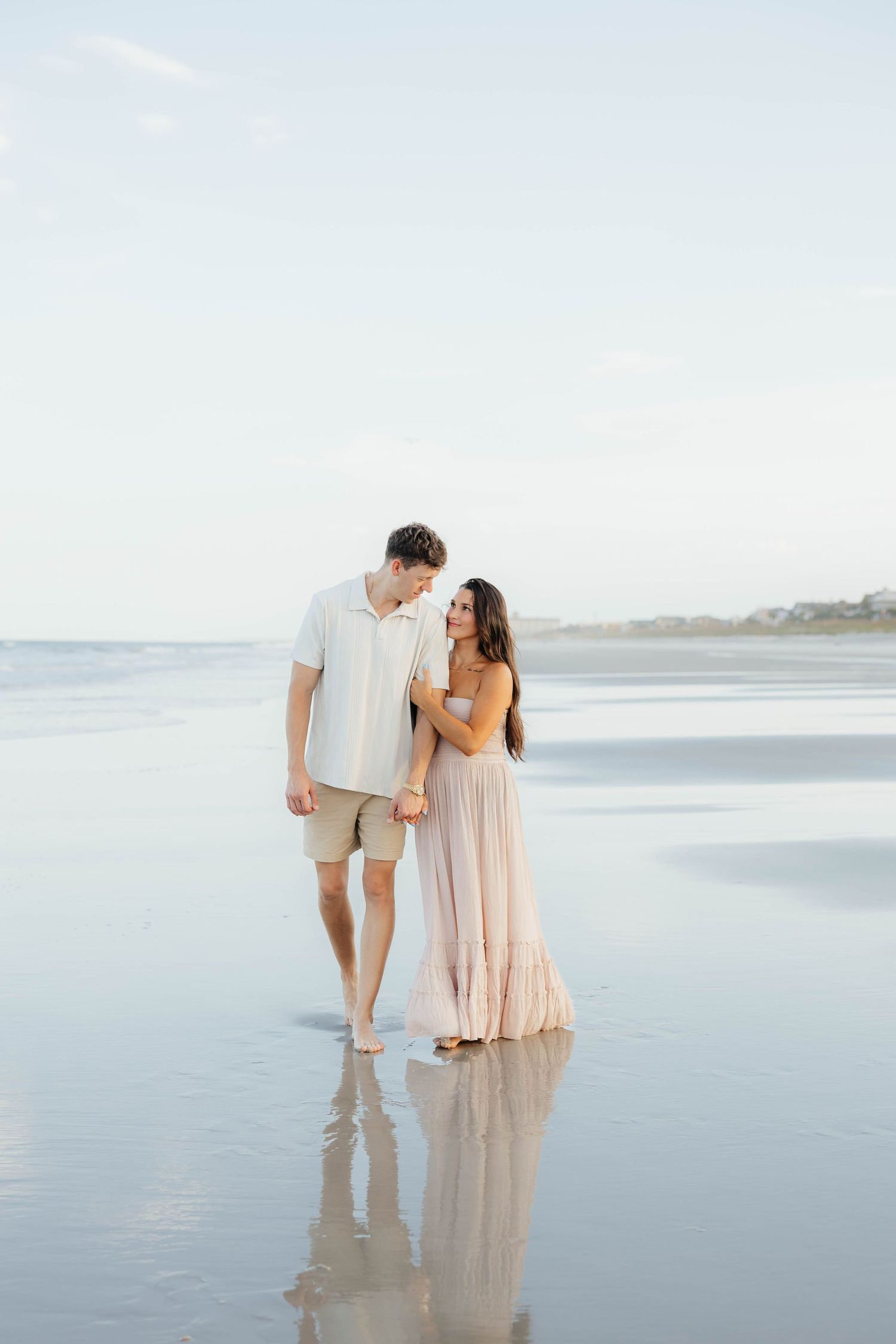 A romantic couple walks together on a reflective beach at sunset wearing light colored clothing.