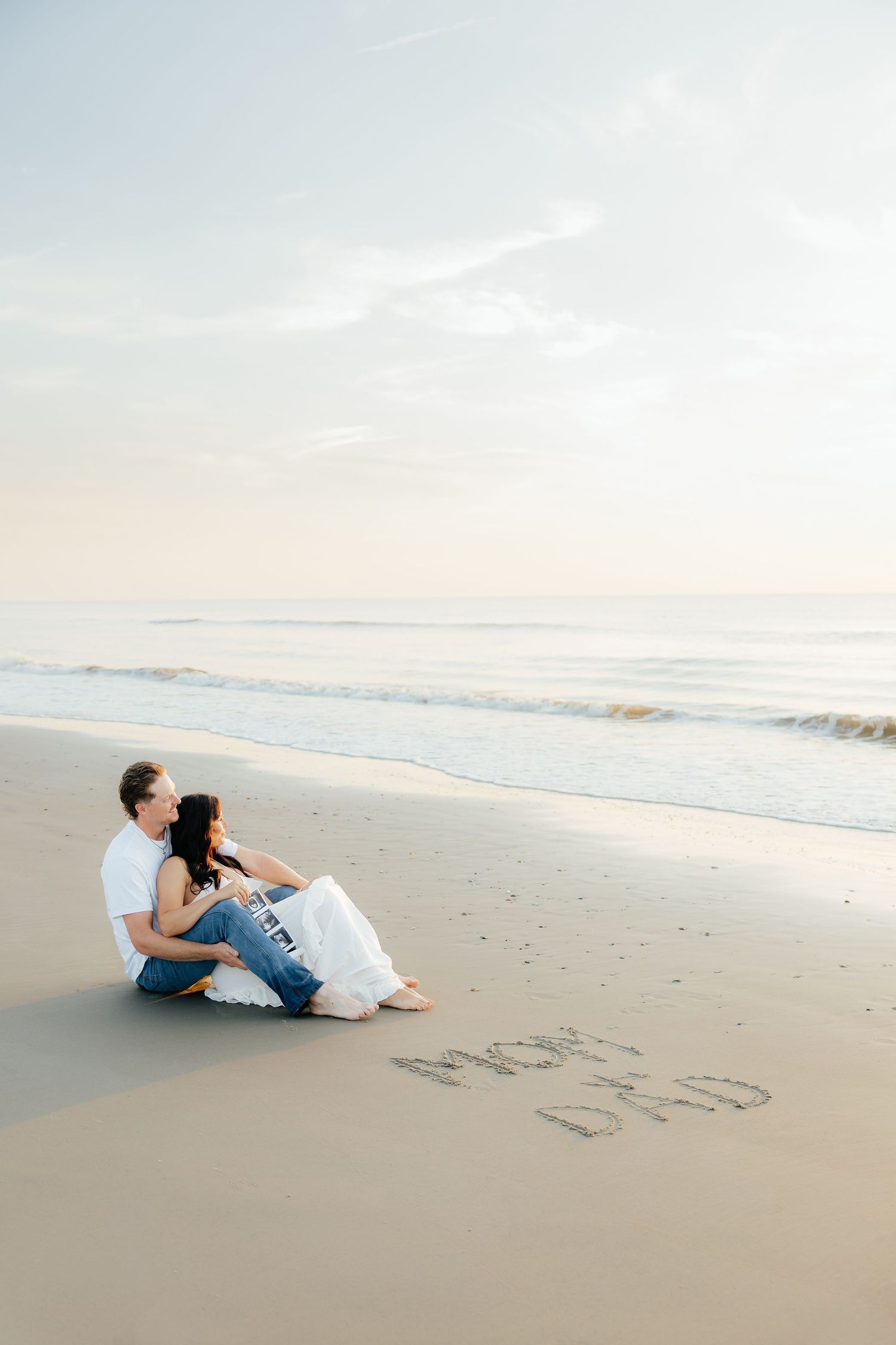 Couple sitting on beach sand with 'Mom & Dad' written in sand, ocean waves in background at sunset.