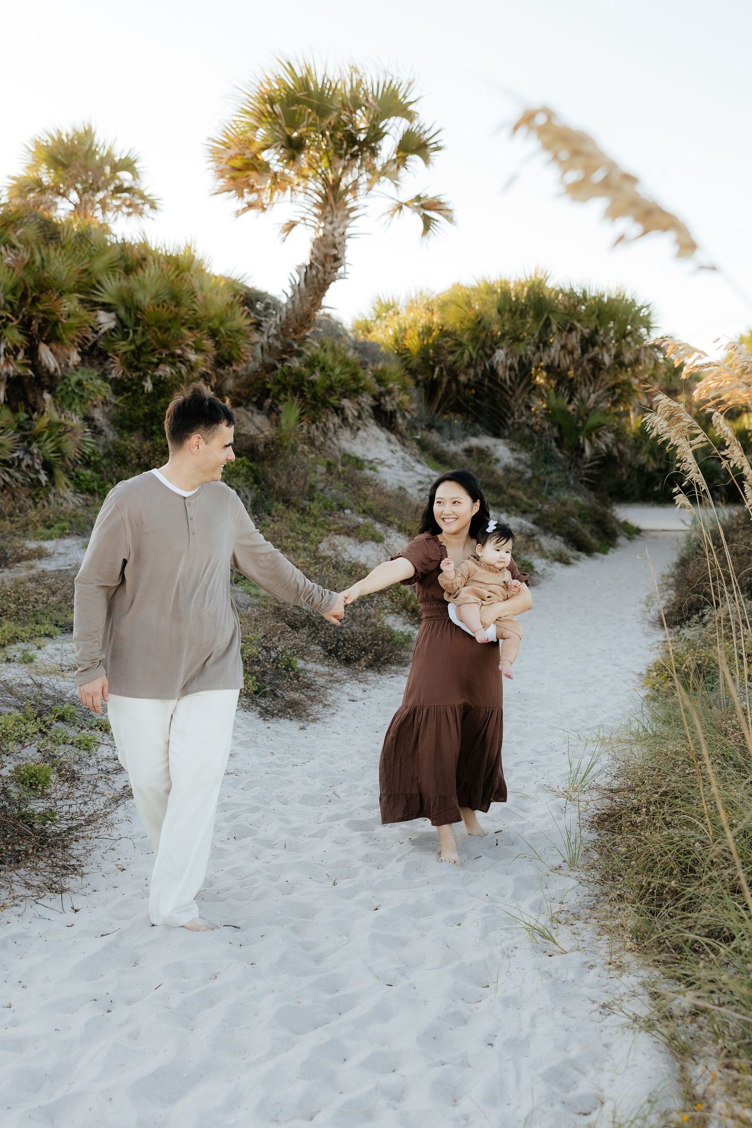 A couple stands together on a sandy beach path surrounded by palm trees and coastal vegetation at sunset.