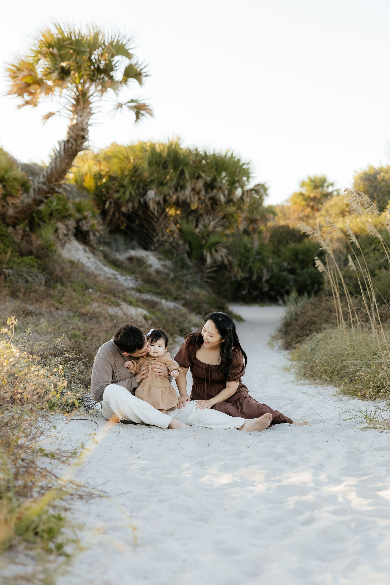 Couple lying together on white sandy beach path surrounded by coastal vegetation at sunset.