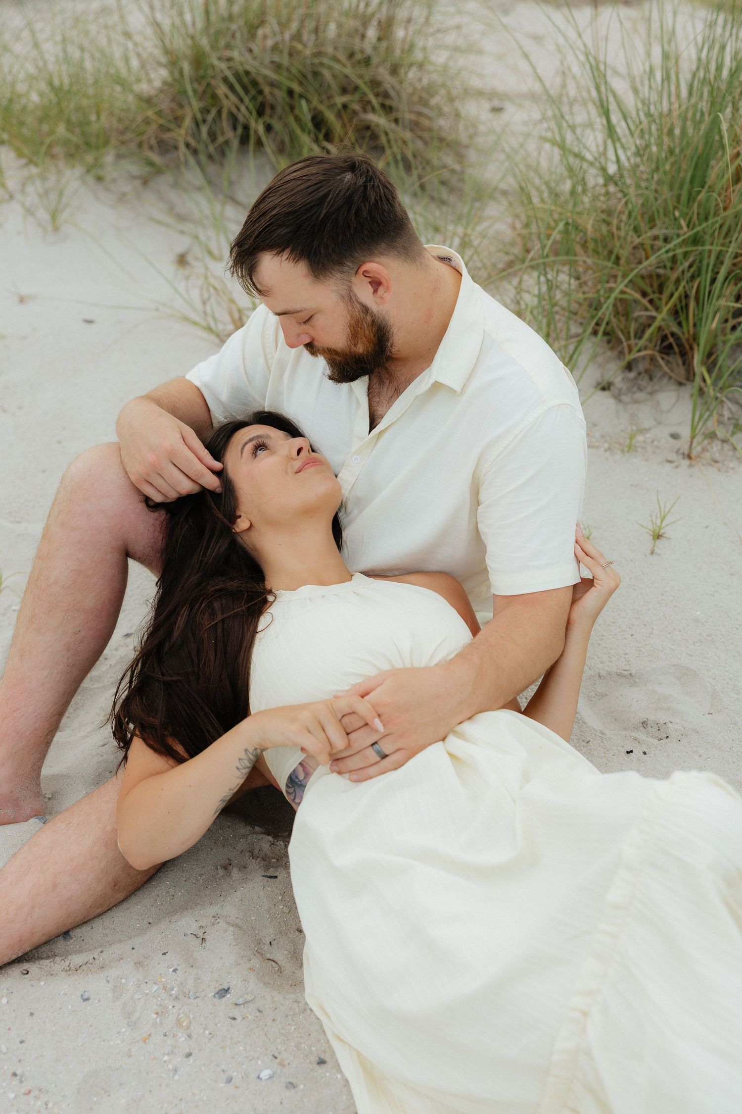 A romantic couple in white outfits embrace on a sandy beach with sea grass in the background.
