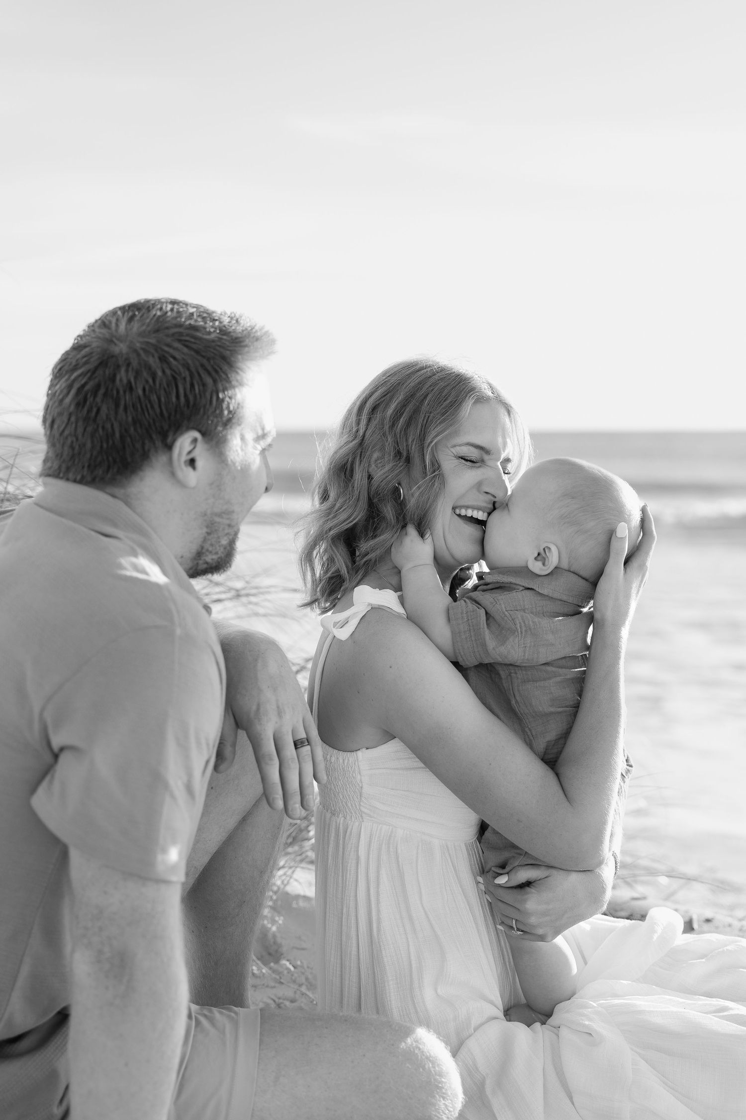 Black and white beach family portrait of a smiling mother cuddling her baby while the father looks on, seated on the sand by the ocean.