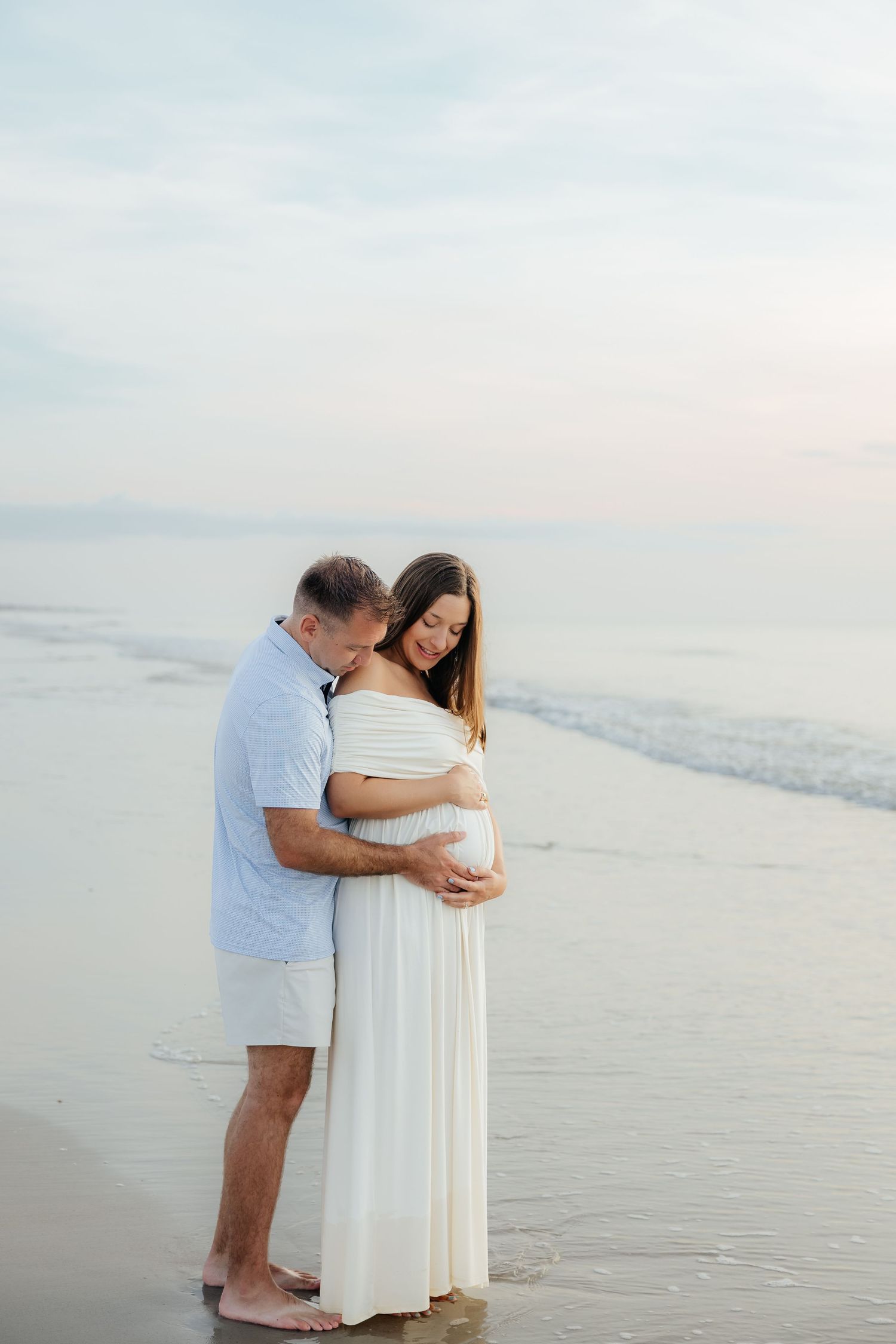 Couple embracing on beach at sunset, pregnant woman in white dress with partner in blue shirt, maternity photo.