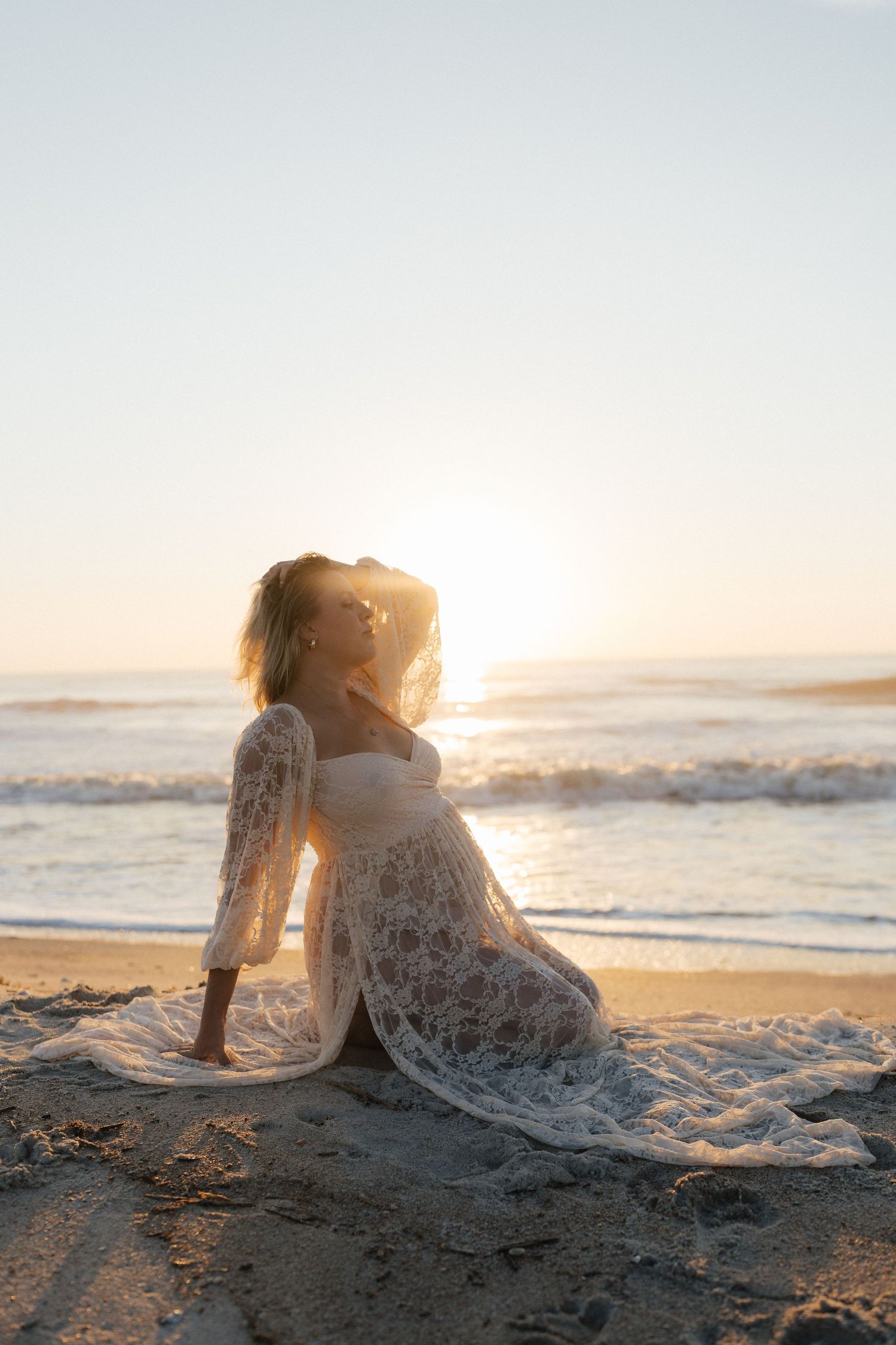 Silhouette of woman in lace dress sitting on beach during golden hour.