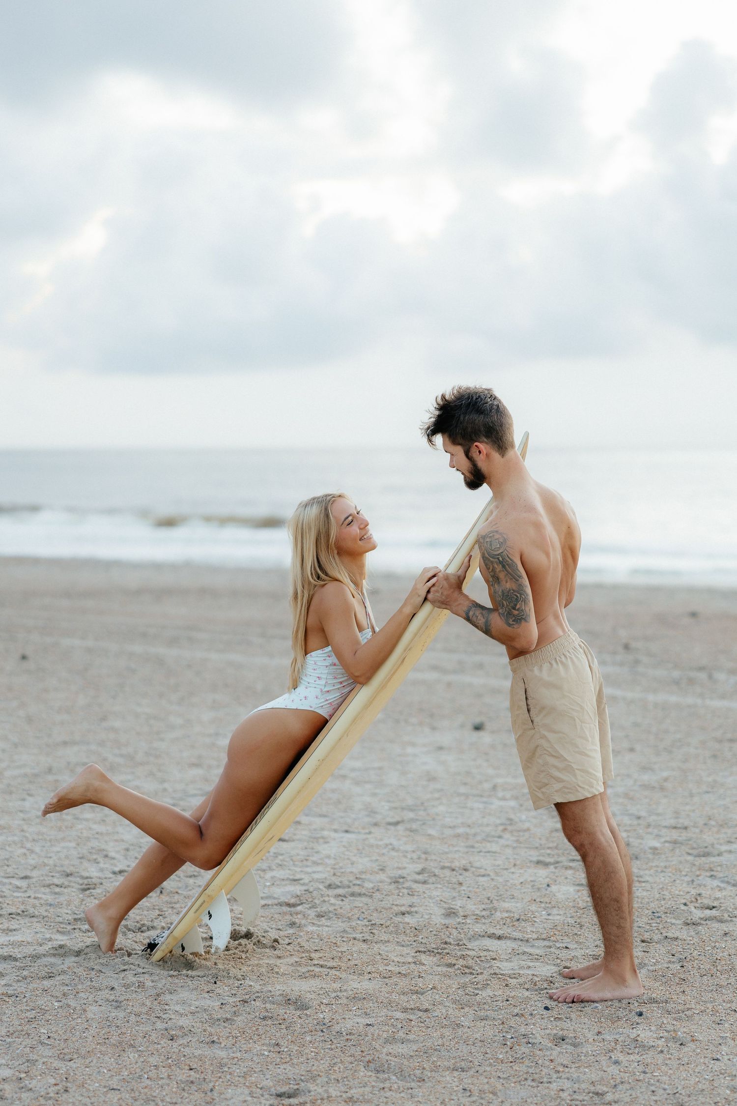 Playful beach couple posing with surfboard on sandy shore.