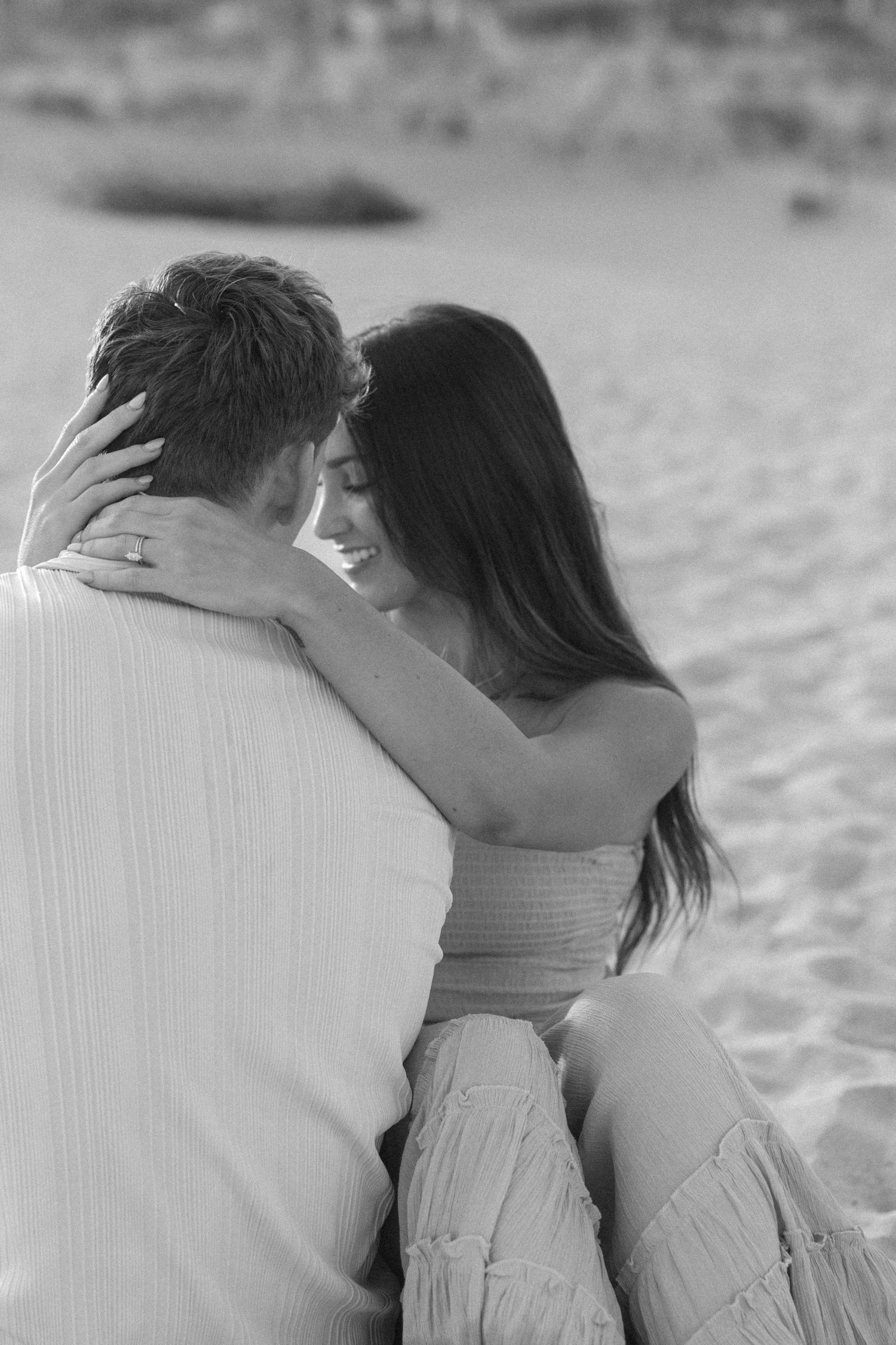 A romantic black and white photo of a couple embracing on a beach, shot from behind.