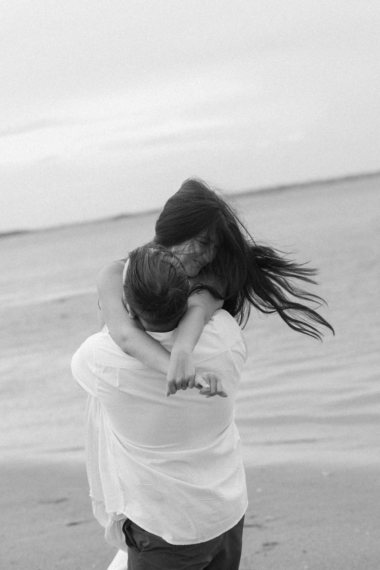 A joyful couple in white clothing embrace and spin on a misty beach in black and white.