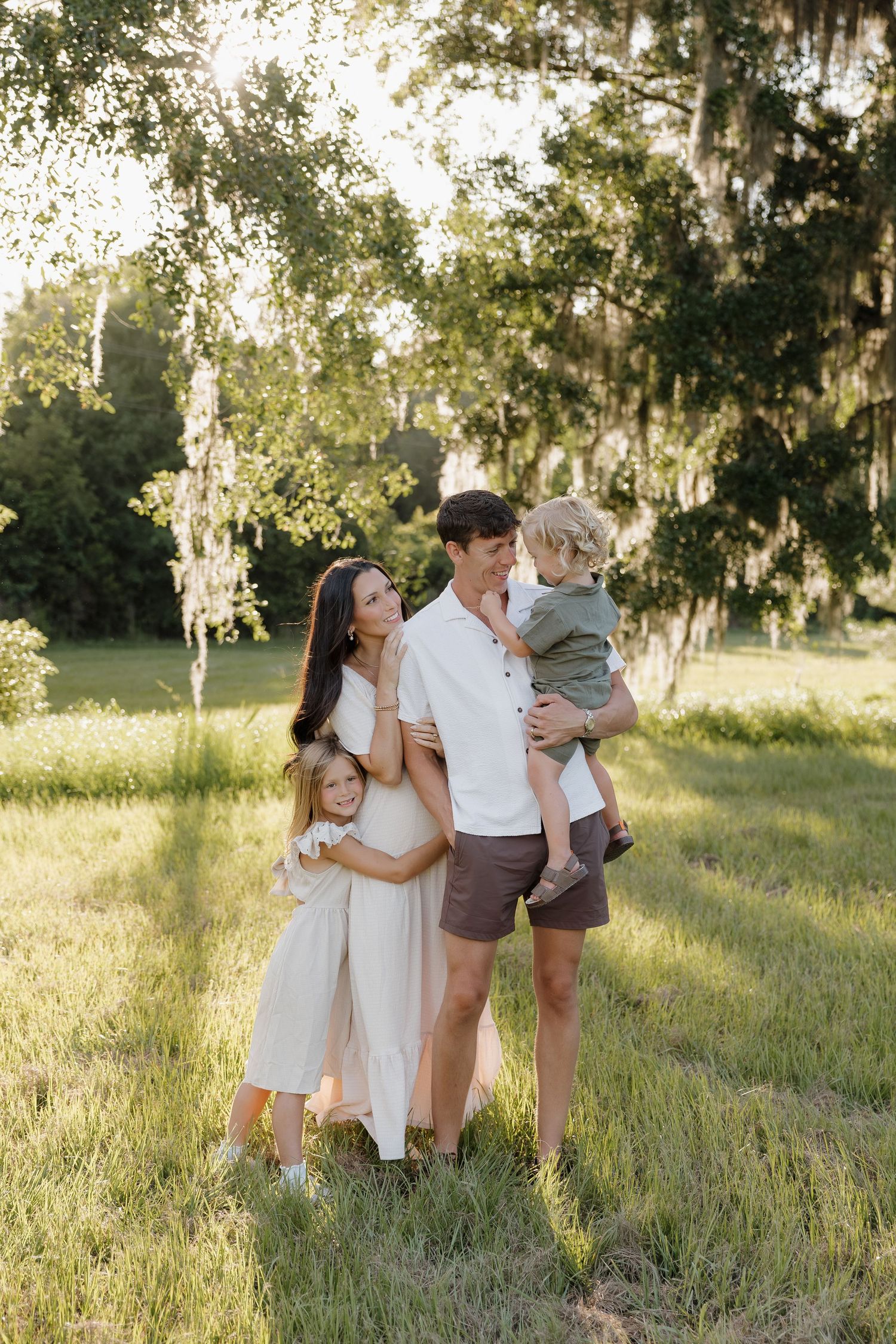 Smiling family of four embracing in a sunlit grassy field beneath oak trees draped with Spanish moss during a golden hour portrait session.