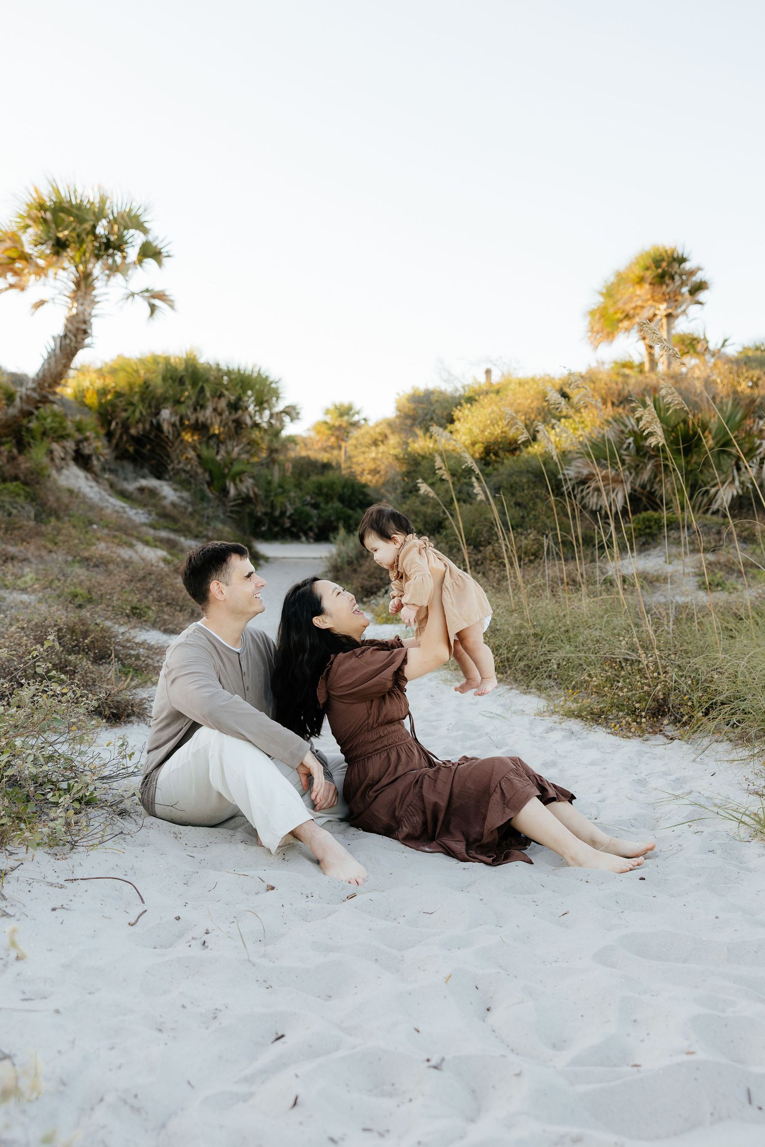 A couple relaxes together on a sandy beach at sunset with trees and greenery in the background.