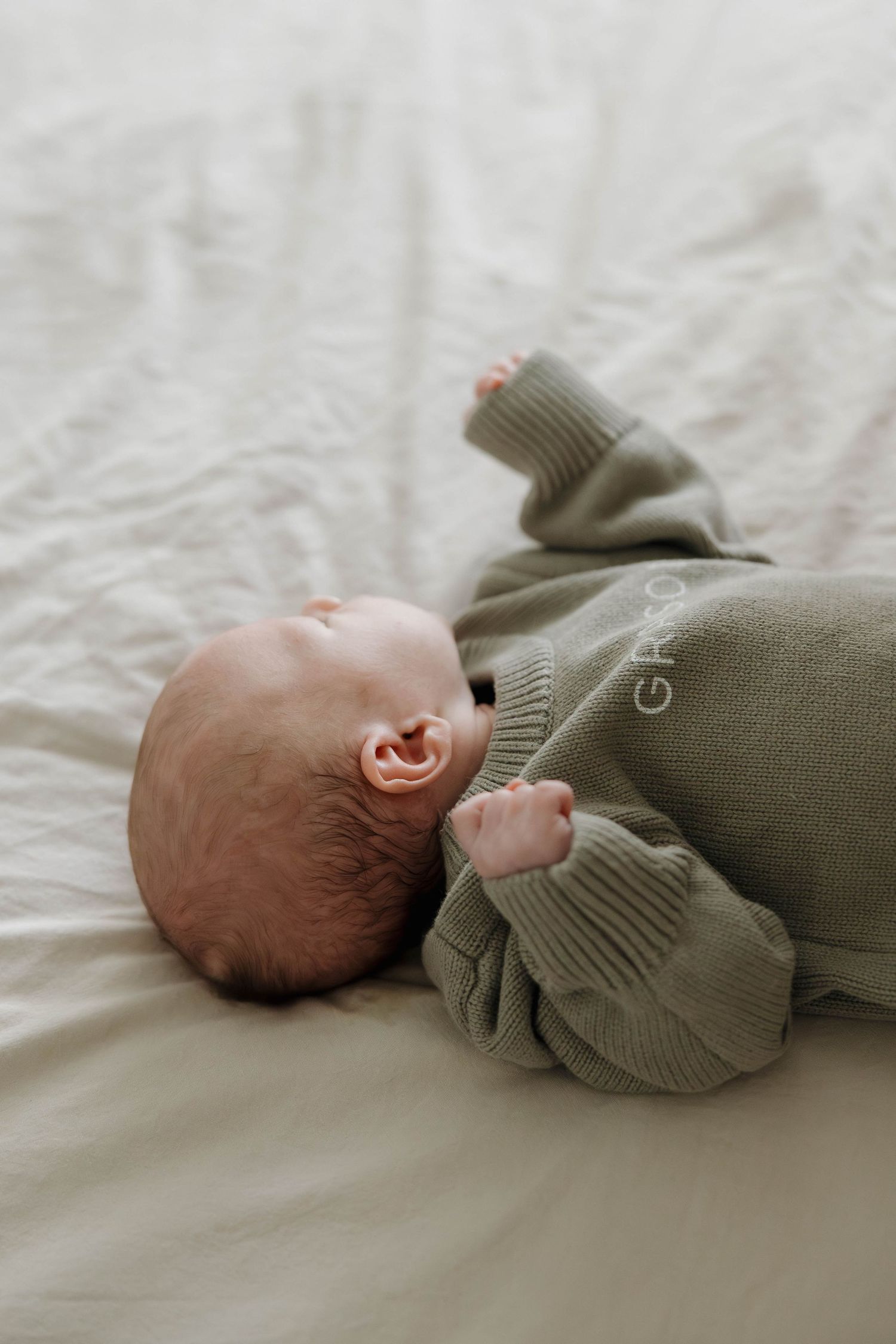 Baby in olive green sweater lying on white bedding in a series of three connected photos.