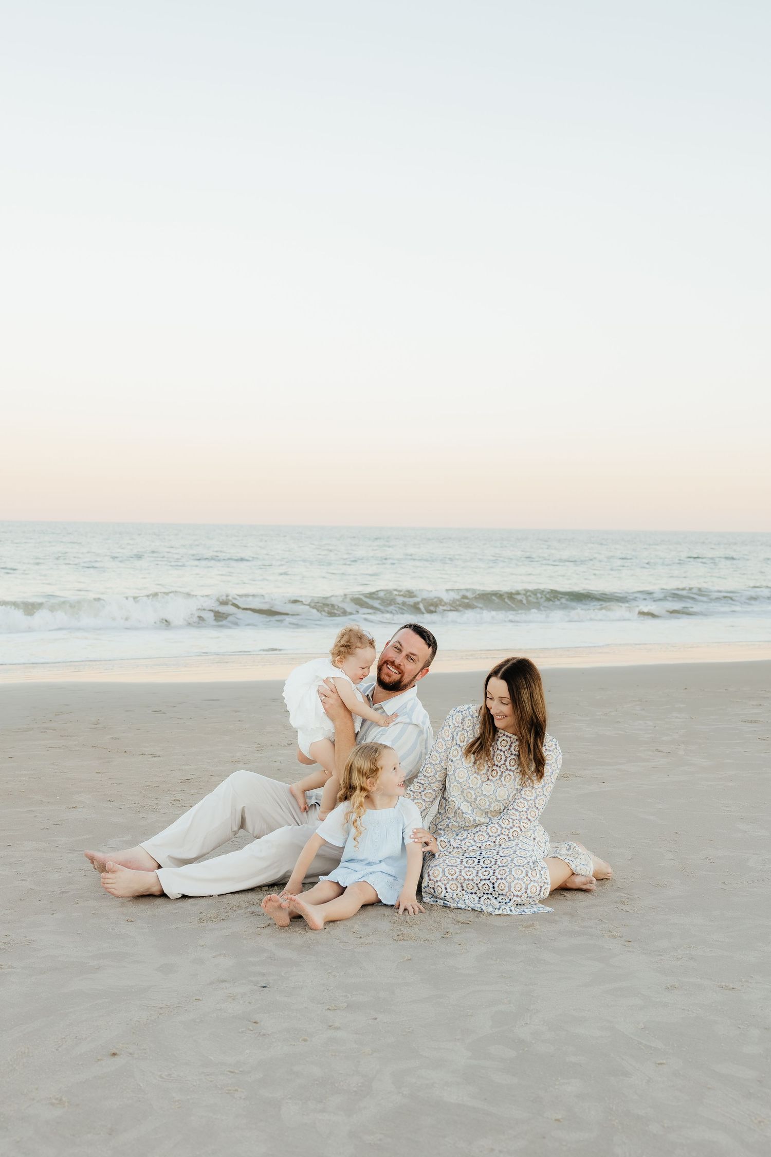 Family of four sitting together on a sandy beach at sunset, with two young children, ocean waves, and a soft pastel sky behind them.