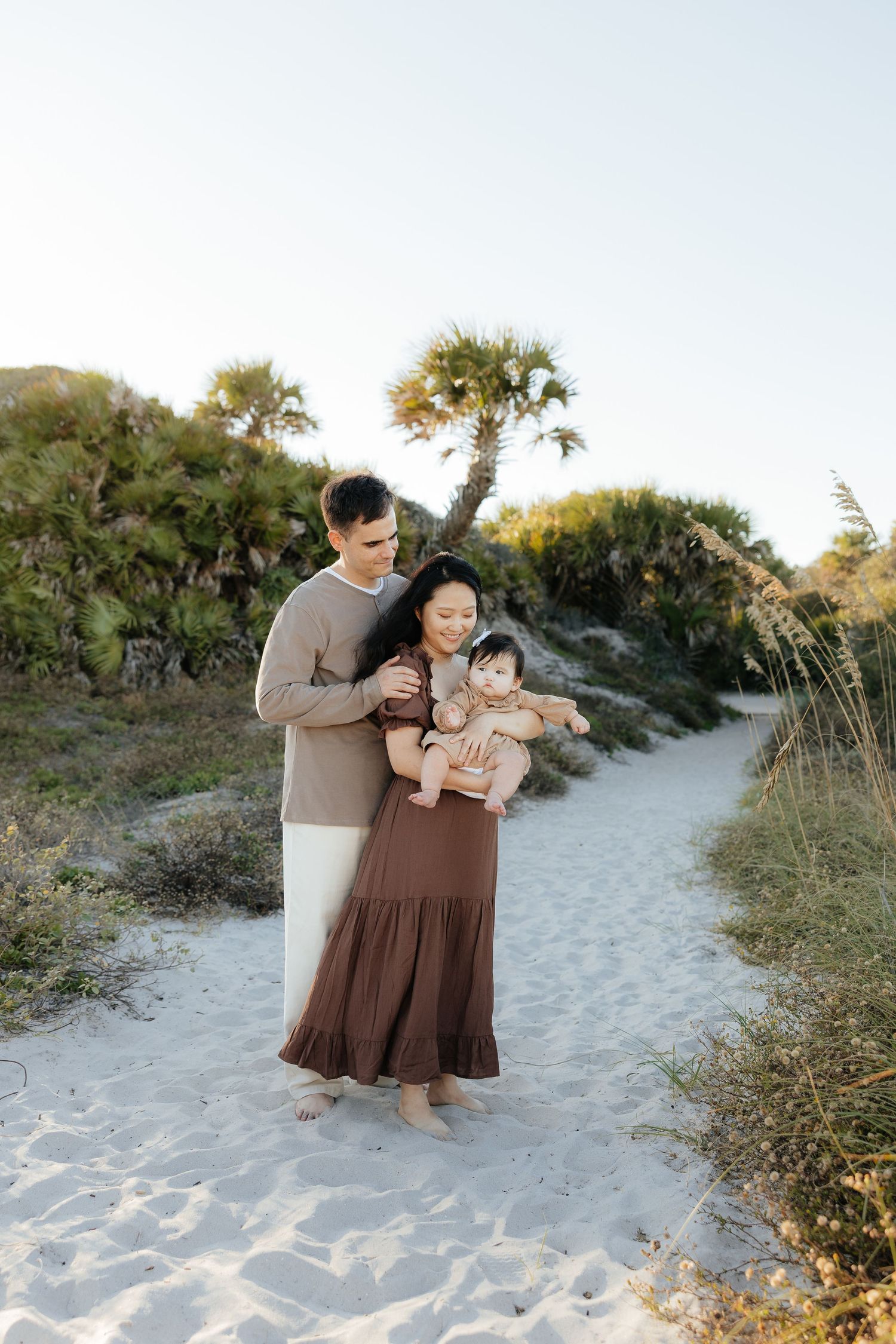 A couple and their baby pose together on a sandy beach path at sunset surrounded by dunes and vegetation.