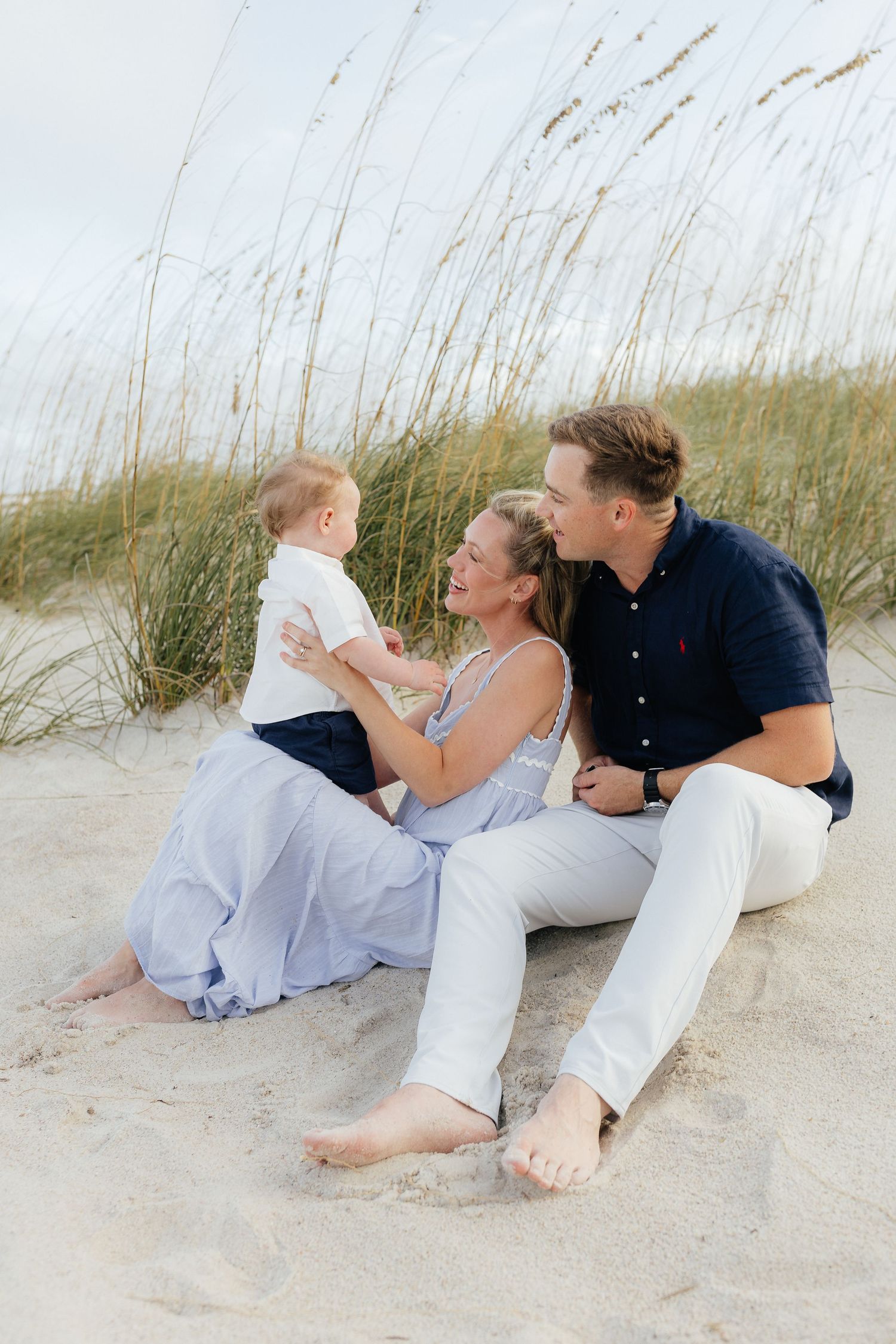 Family relaxing together on sandy beach dunes with coastal grass in background.