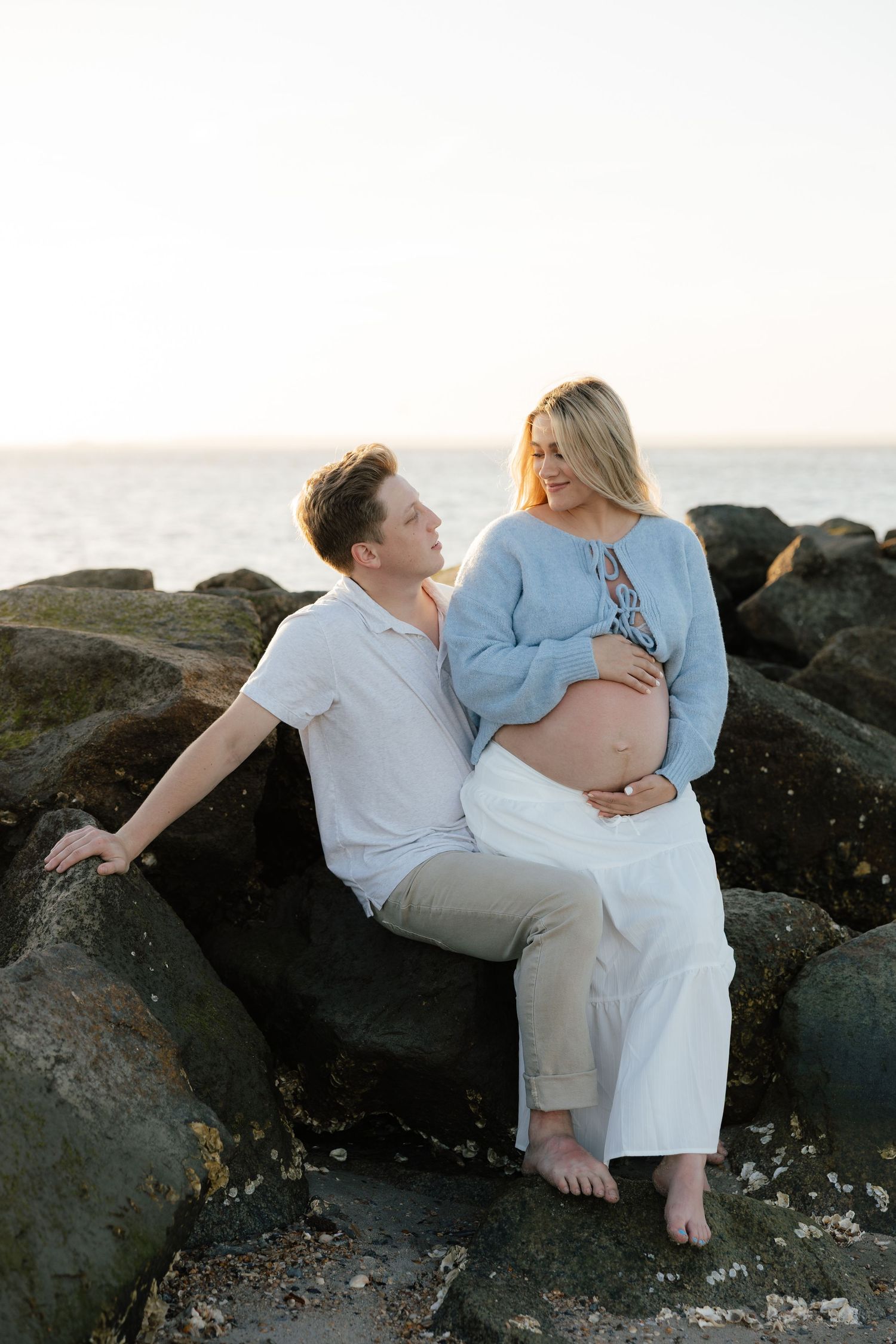 Couple poses together on rocky beach during golden hour maternity photography session.