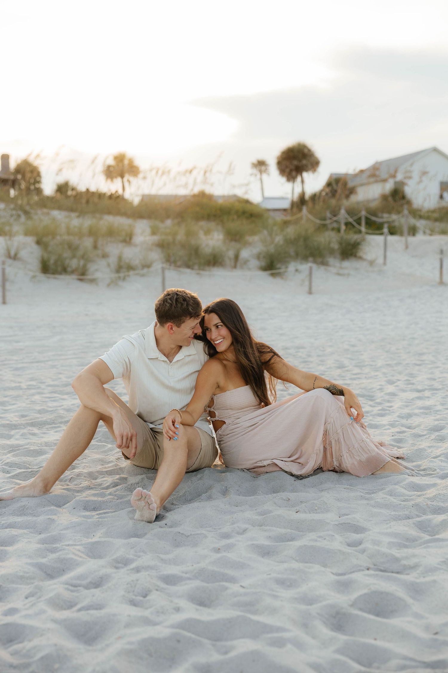 A couple in light colored outfits embraces romantically while laying in soft white sand at sunset on the beach.