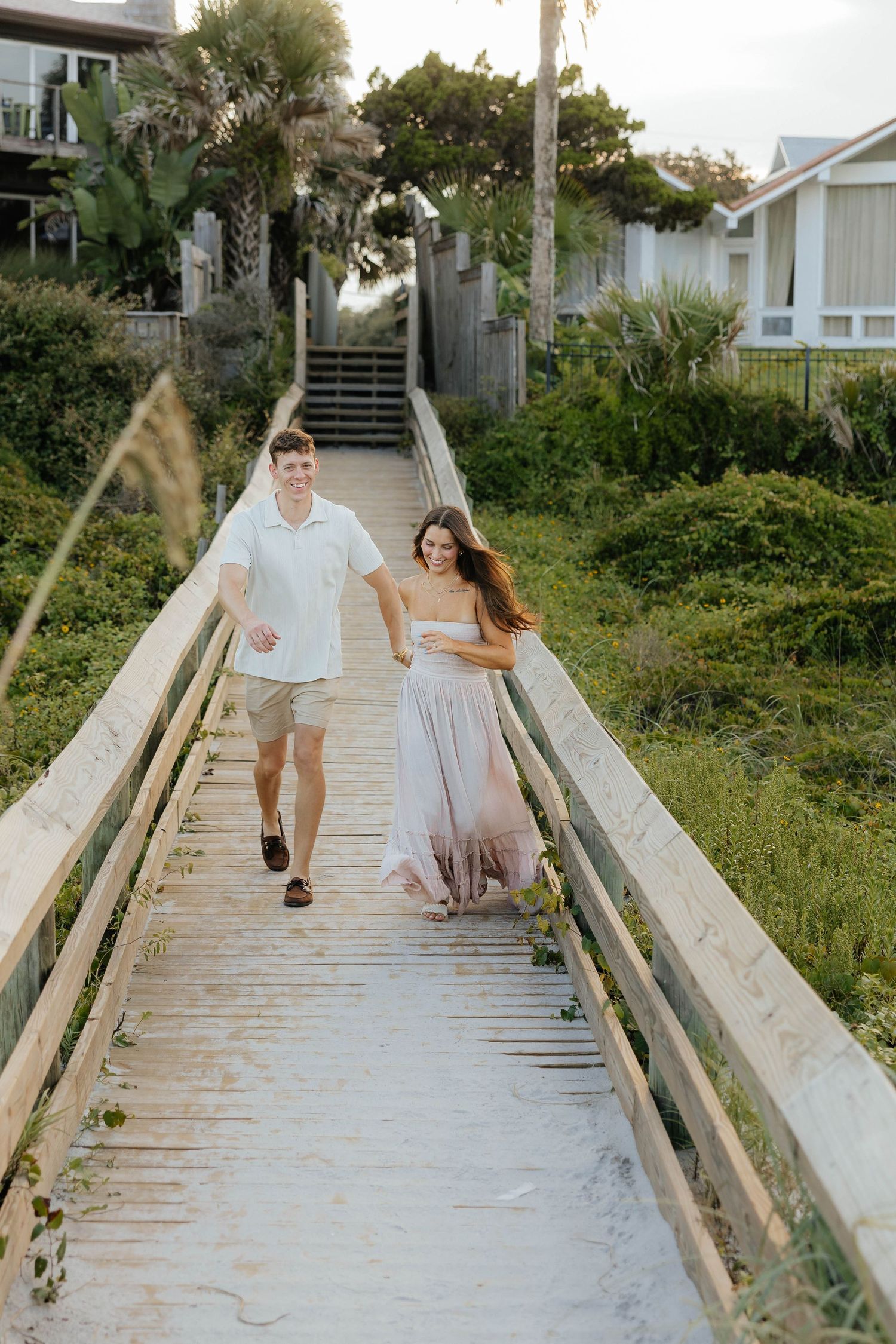 A couple walks hand in hand down a sunlit wooden boardwalk path leading to the beach.