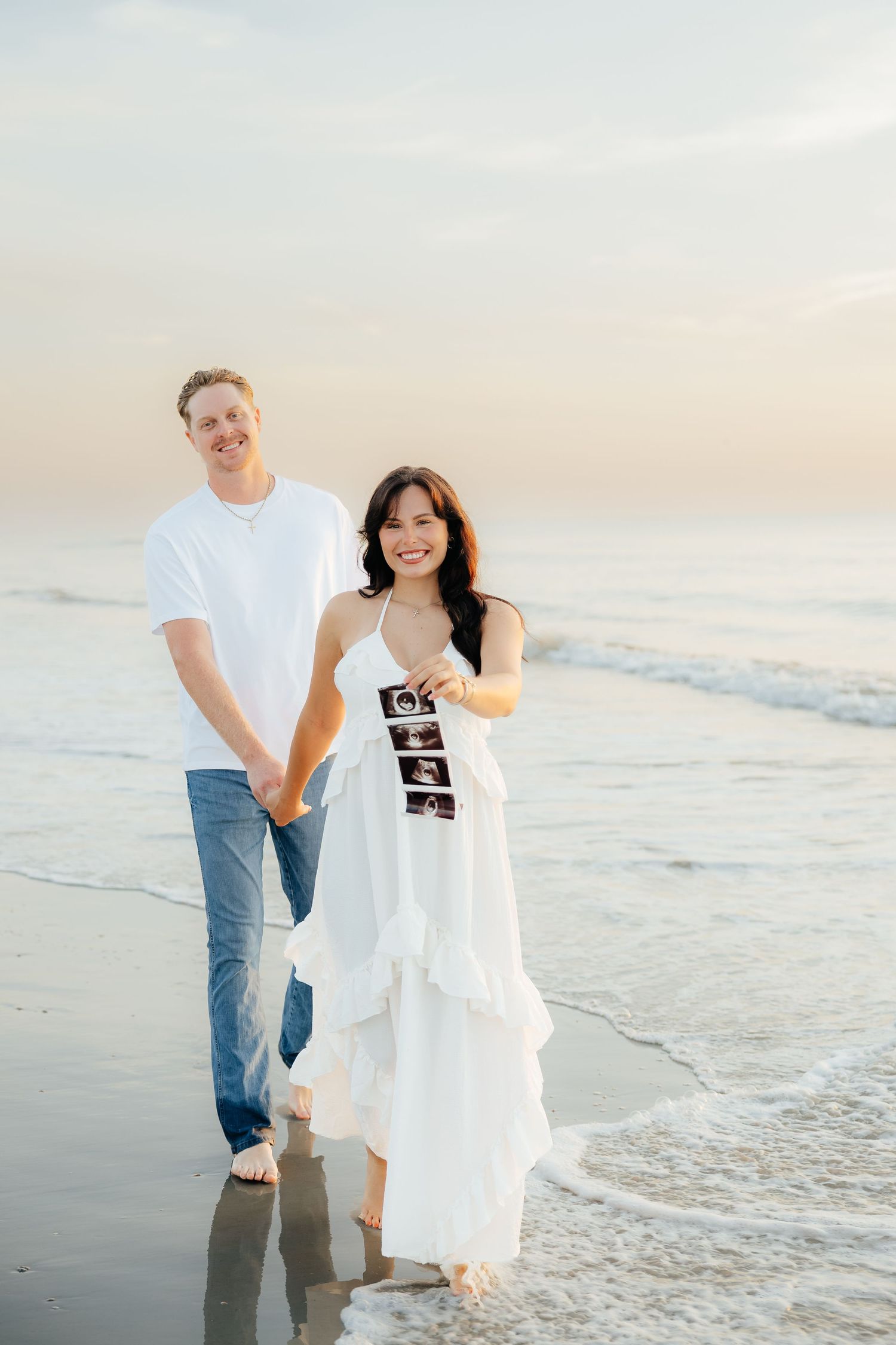 A smiling couple walks hand-in-hand on the beach, the woman in a white dress holding ultrasound photos for a pregnancy announcement.