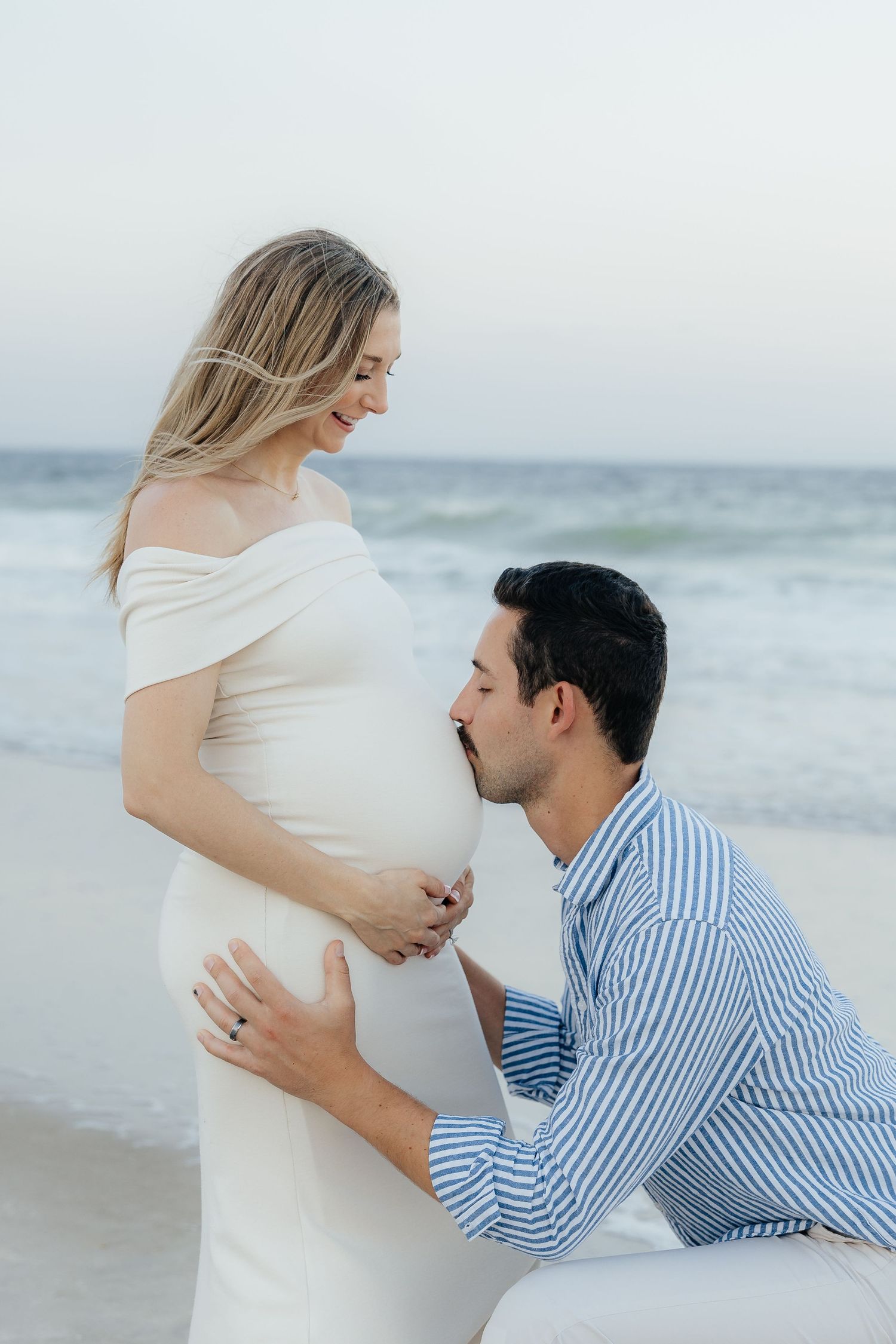 A romantic maternity photoshoot on a beach with a couple in white and blue striped outfits by the ocean waves.