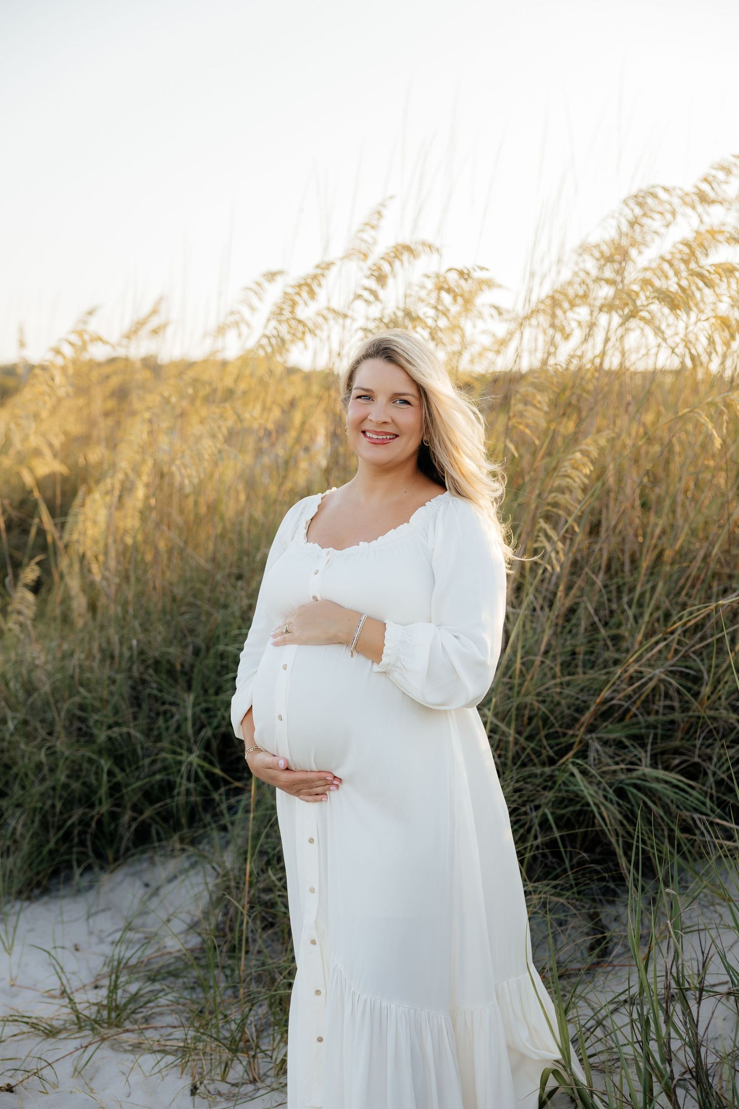Smiling pregnant woman in a white button-front dress cradles her baby bump among golden sea oats on a sandy beach dune at sunset.