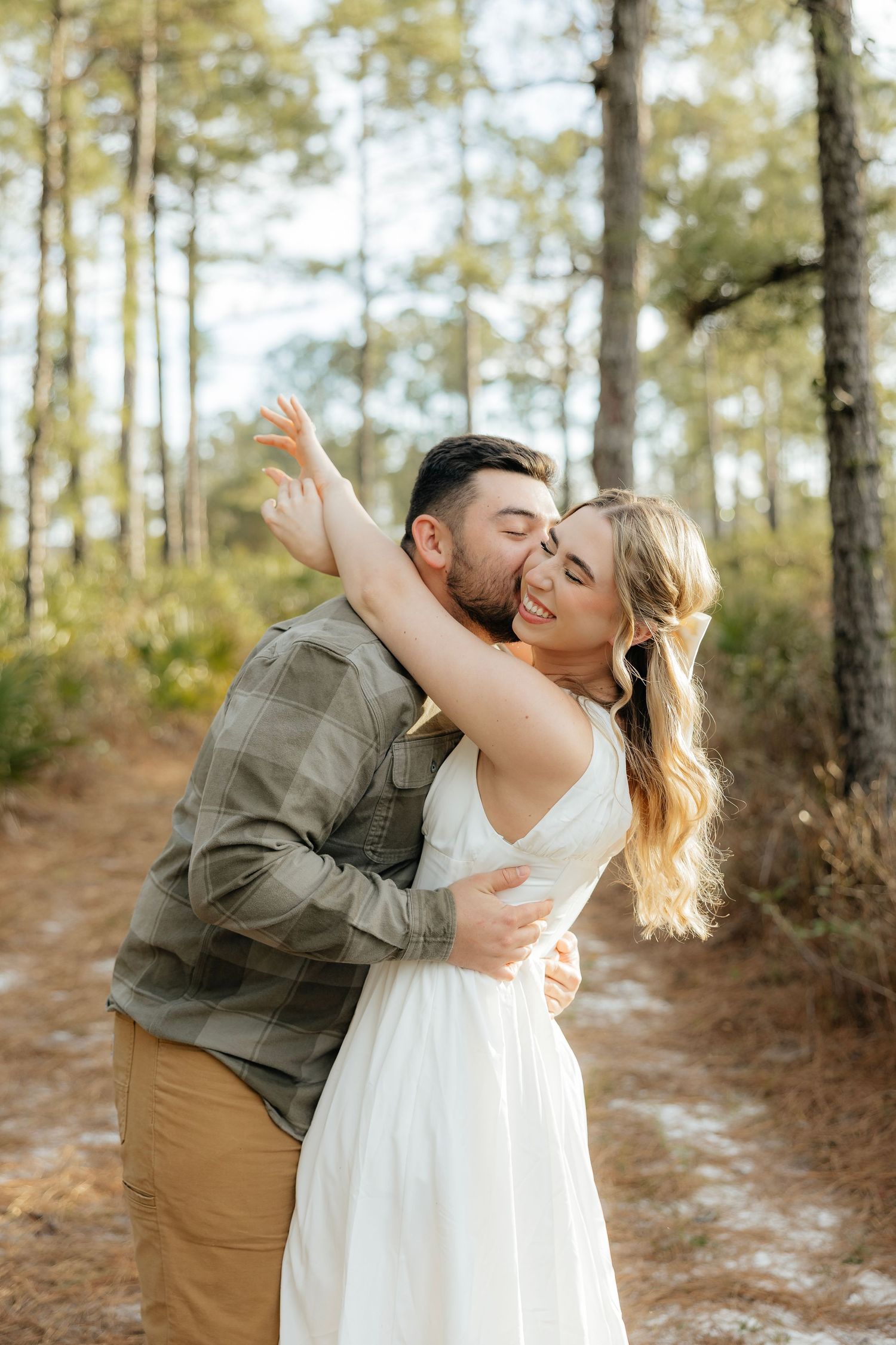 Smiling couple in a loving embrace on a sandy forest path surrounded by tall pine trees, perfect for engagement or couples photography.