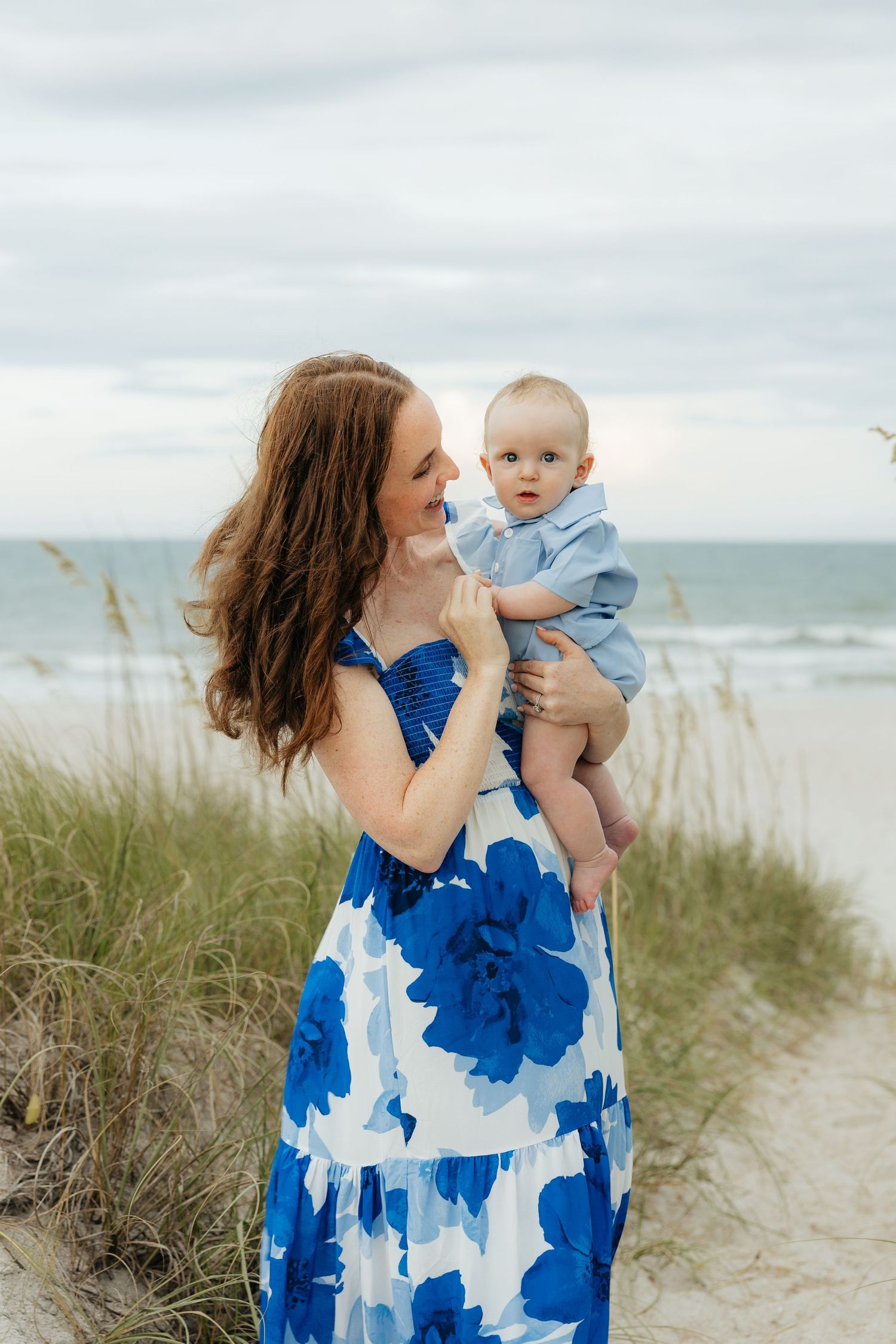 A woman in a blue and white floral dress holds a baby against a coastal beach backdrop with sea grass.
