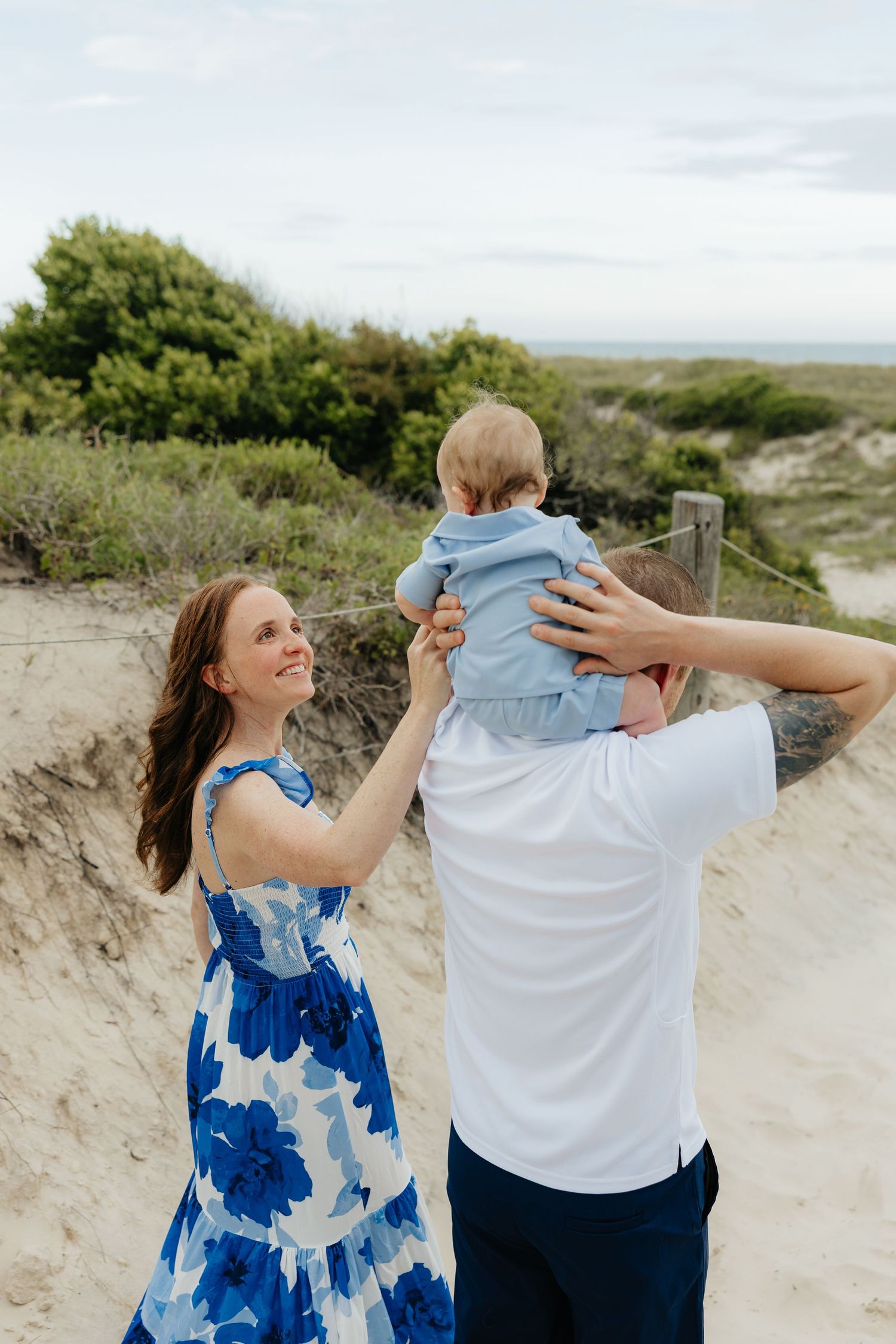 A couple holds their baby on a sandy beach path with dunes and greenery in the background.