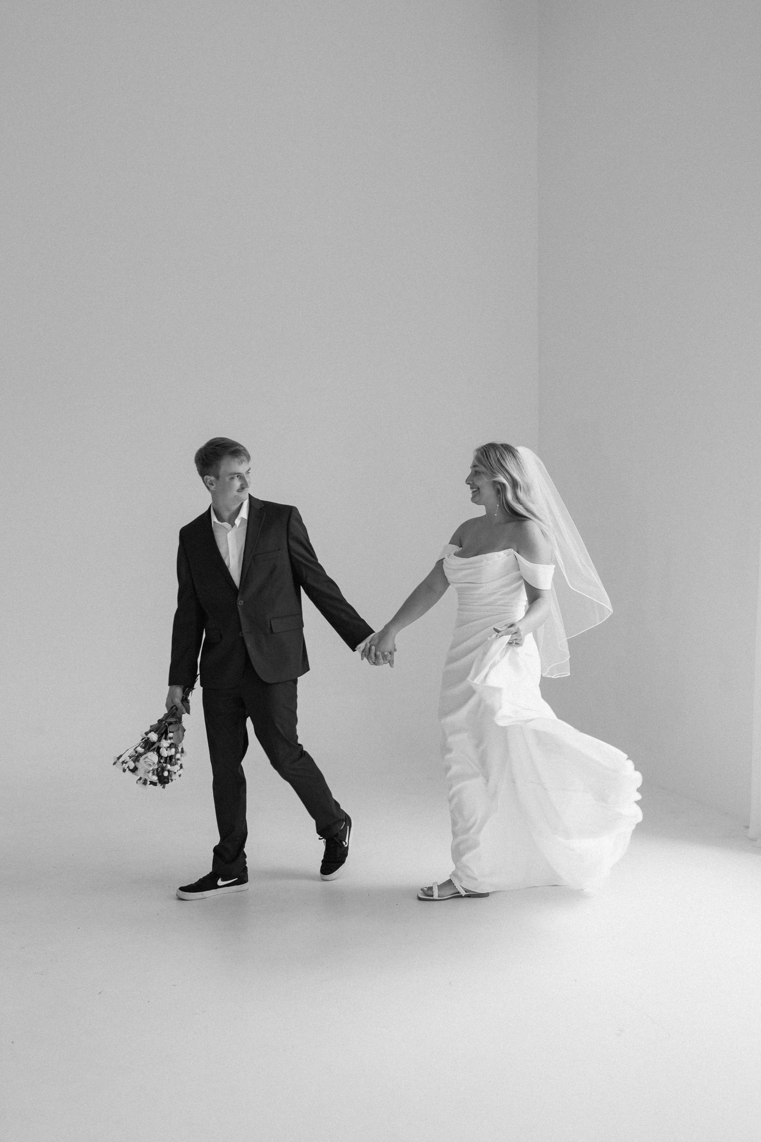 A playful couple in wedding attire dance together against a minimalist white studio background.