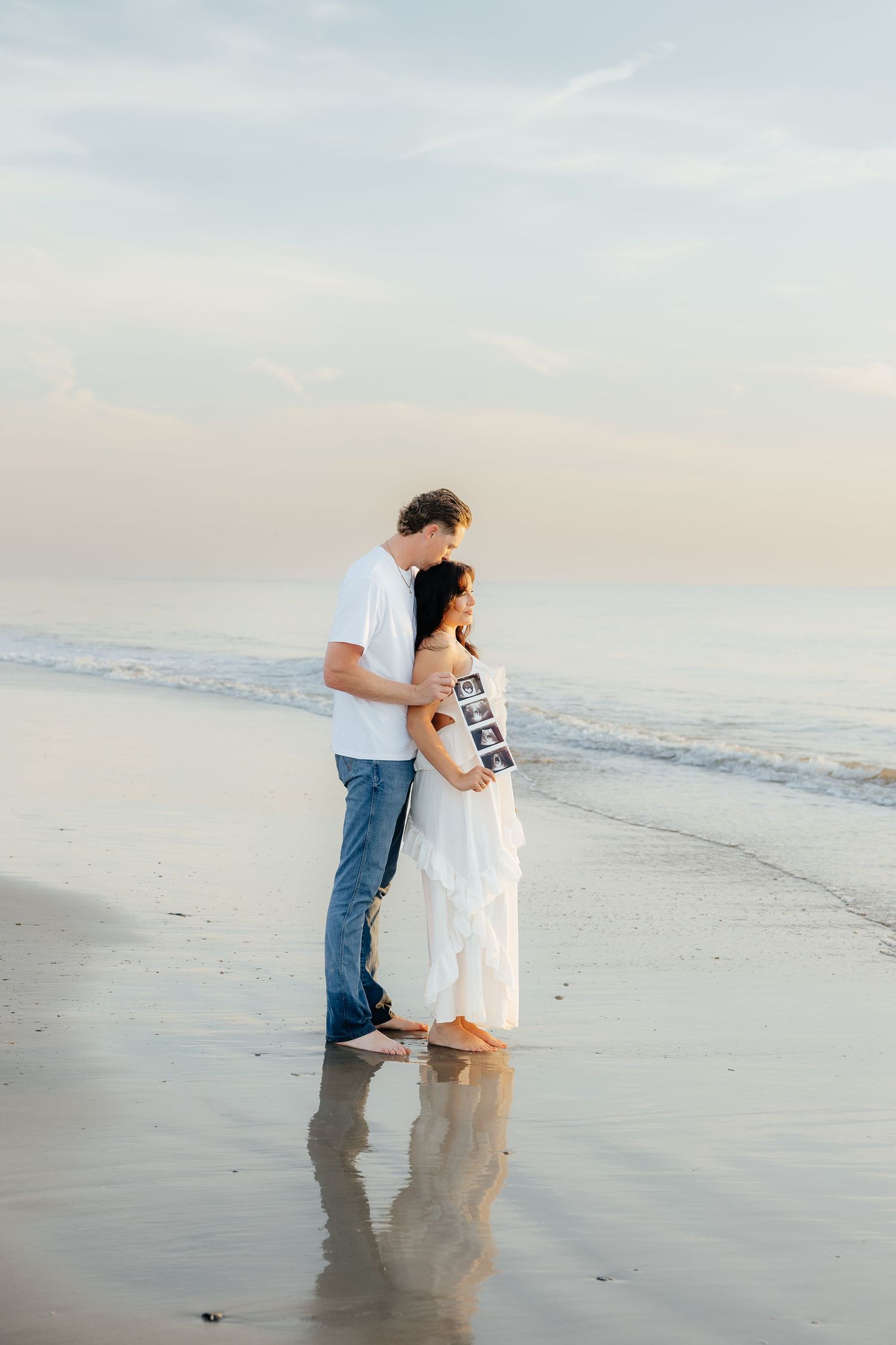 Couple standing on beach holding ultrasound photos, woman in white dress, man in white shirt and jeans, ocean backdrop.