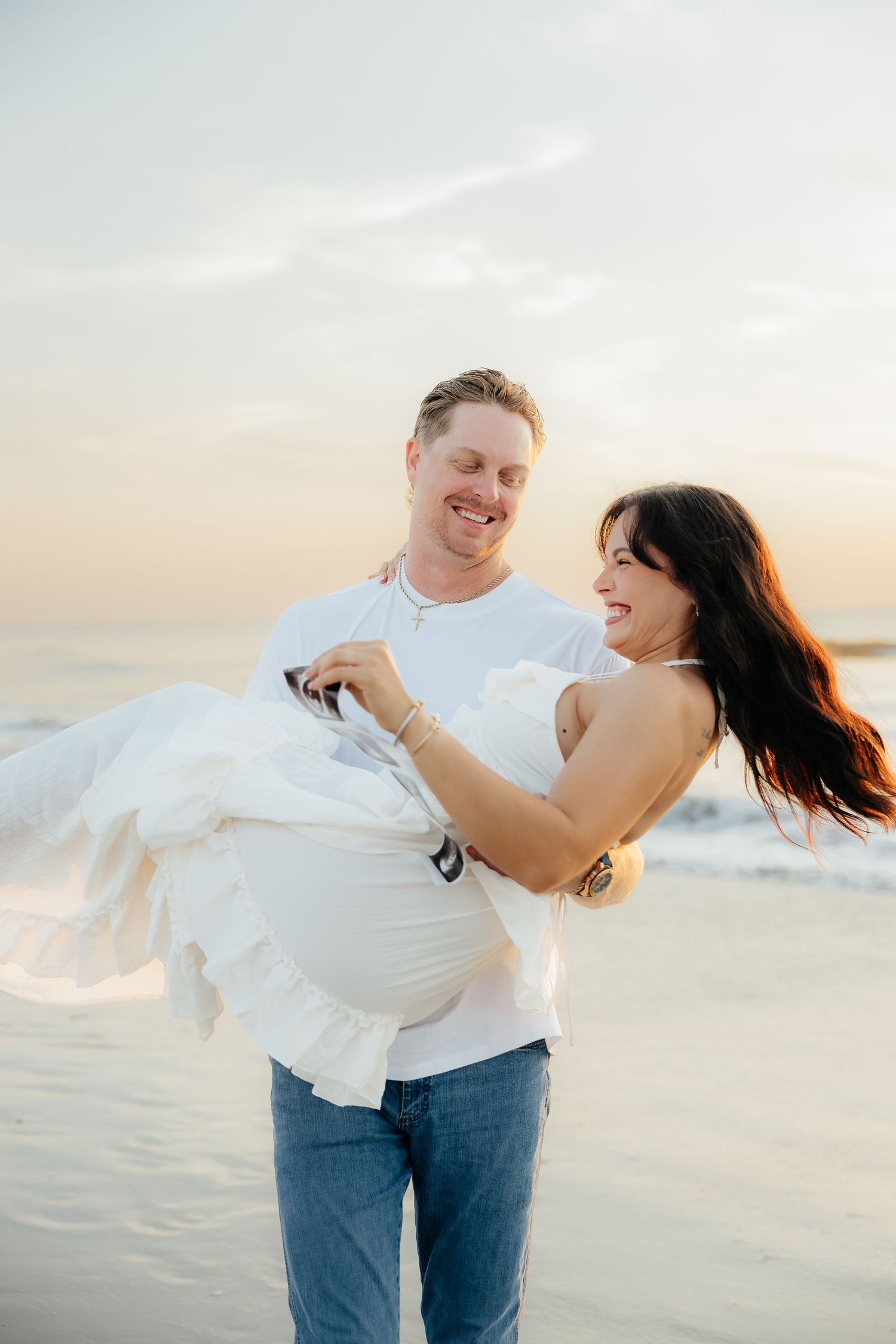 A smiling couple poses on a beach at sunset, with the man carrying his pregnant partner in a flowing white dress.