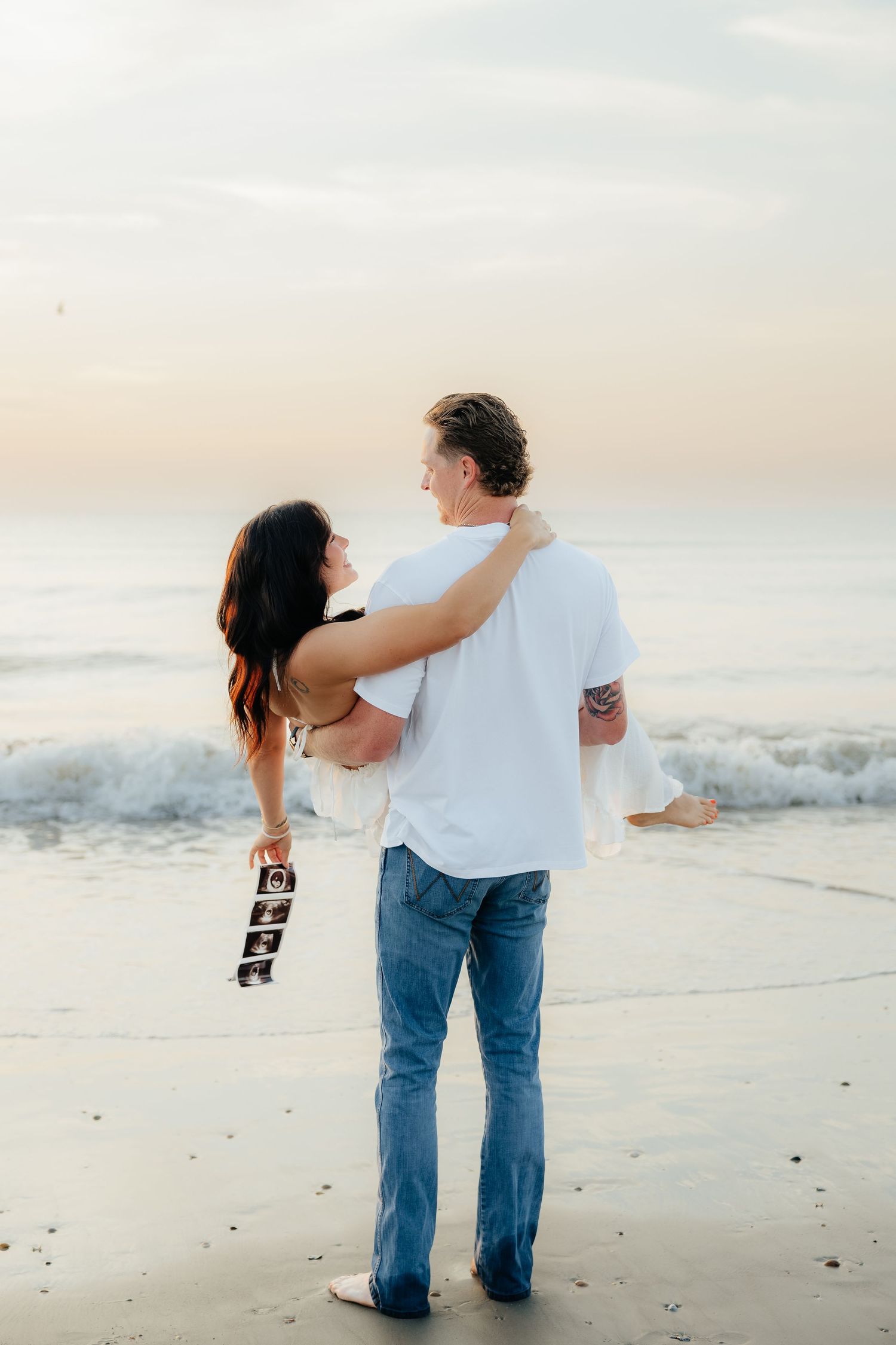 Couple embracing on beach at sunset, woman holding ultrasound photos, announcing pregnancy by ocean waves.