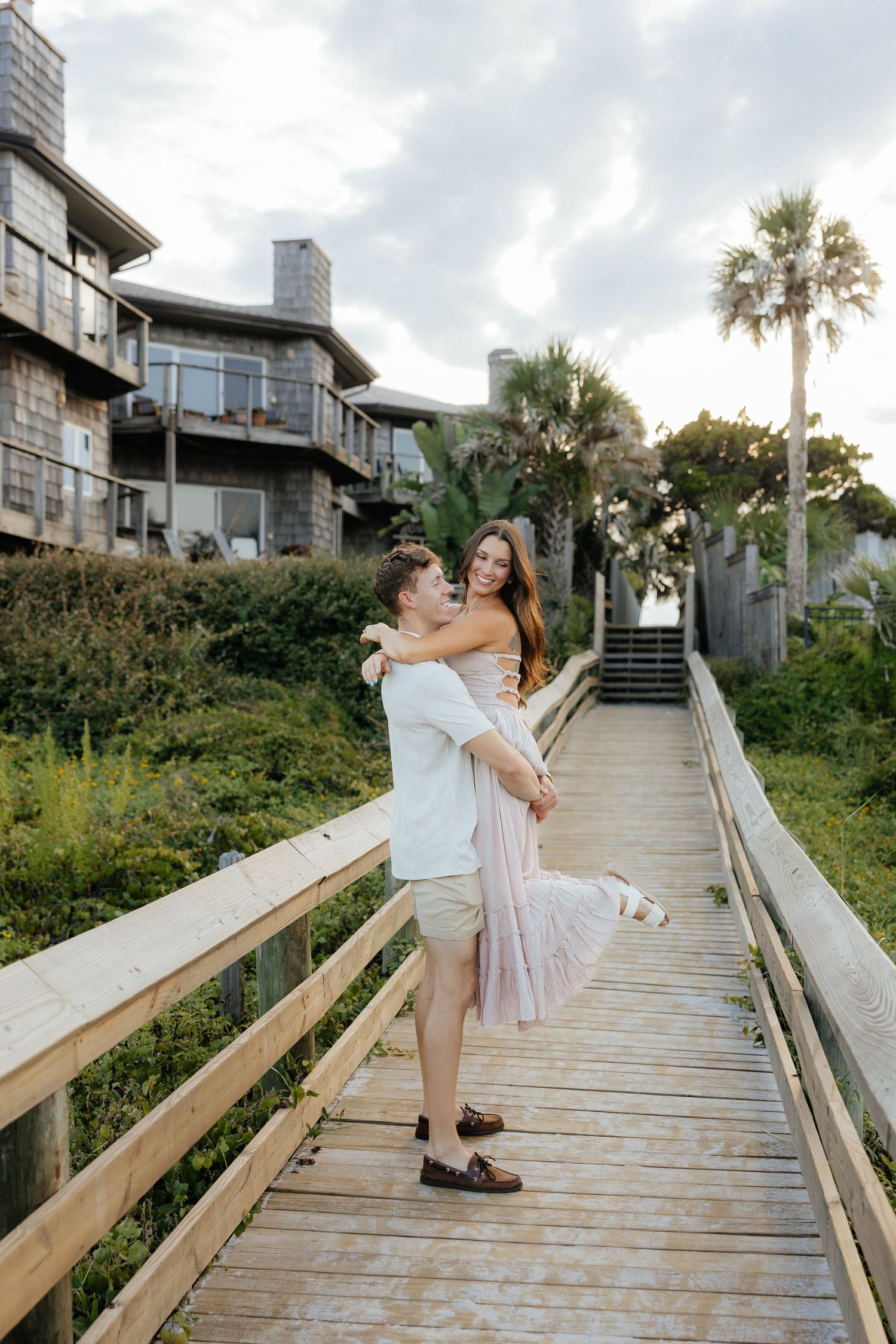 Couple shares romantic moment on wooden beach boardwalk with coastal vegetation and palm trees in the background.