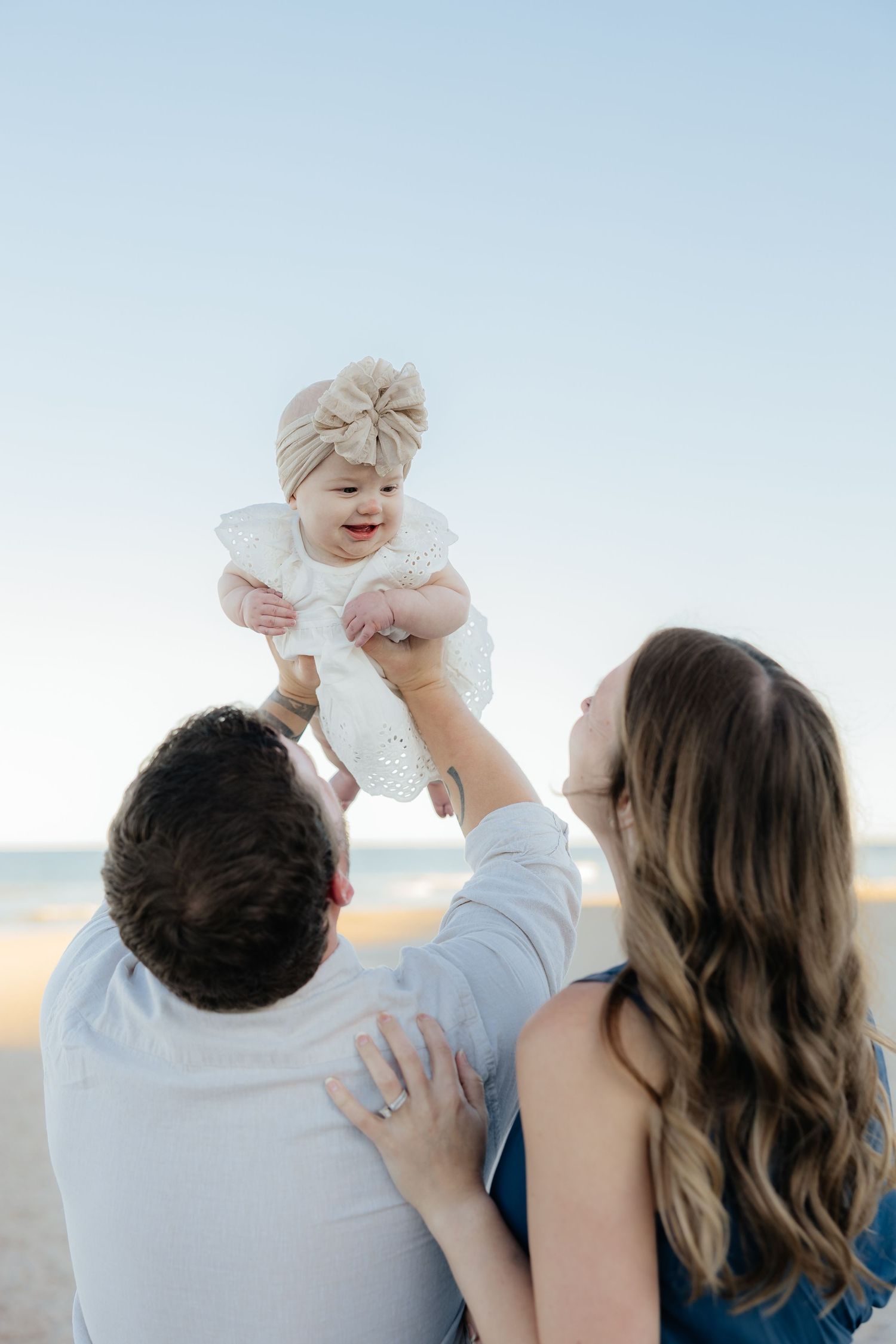 Parents lifting smiling baby girl in white dress and bow headband on a sunny beach.