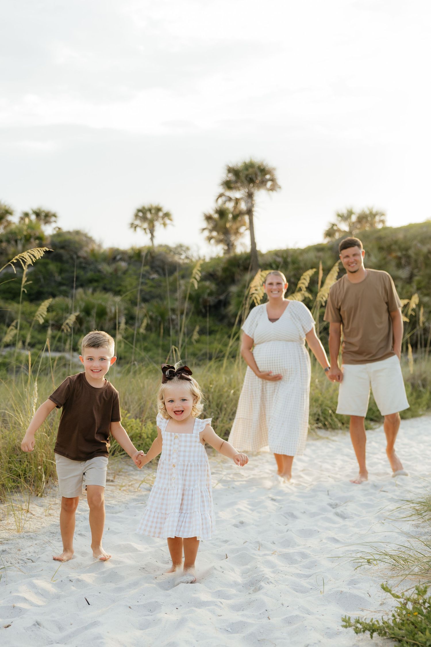 Family of four walking barefoot on a white sand beach path with palm trees and sea oats, mother expecting, during a sunset photoshoot.