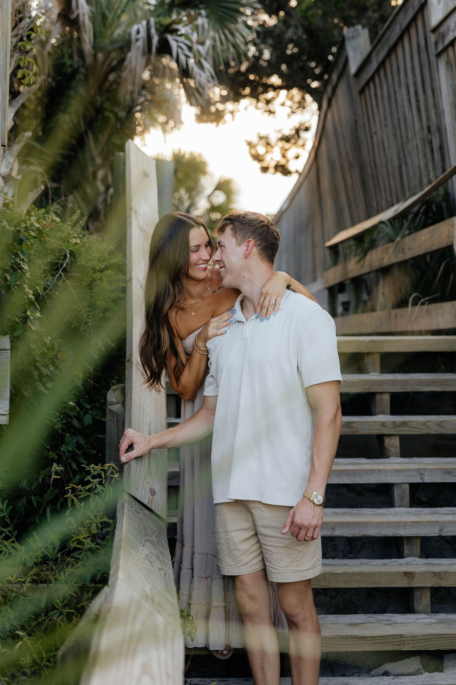 A couple shares romantic moments on a wooden boardwalk surrounded by lush greenery on a sunny afternoon.