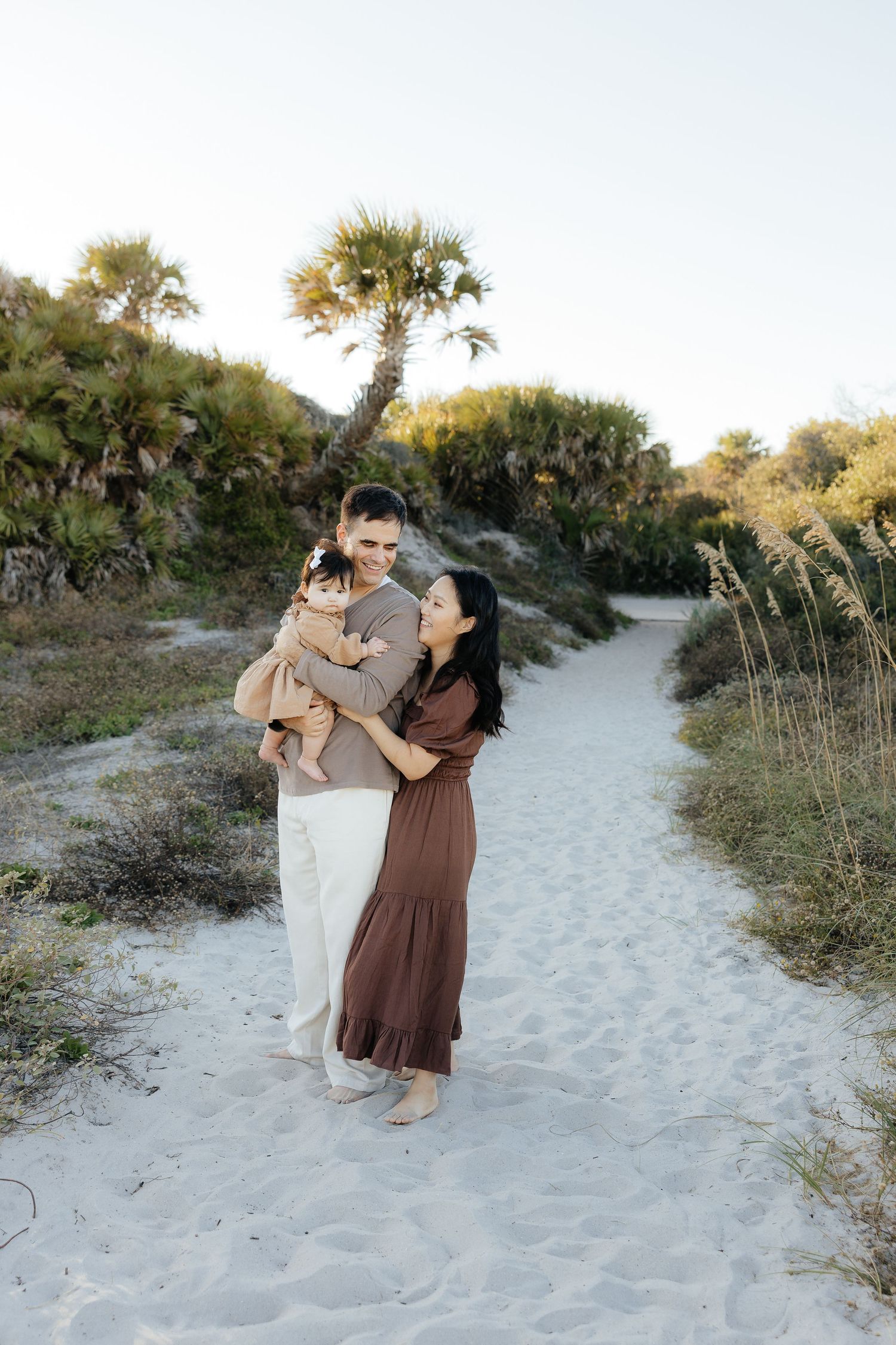 A couple and their baby pose together on a sandy beach path surrounded by palm trees and coastal vegetation at sunset.