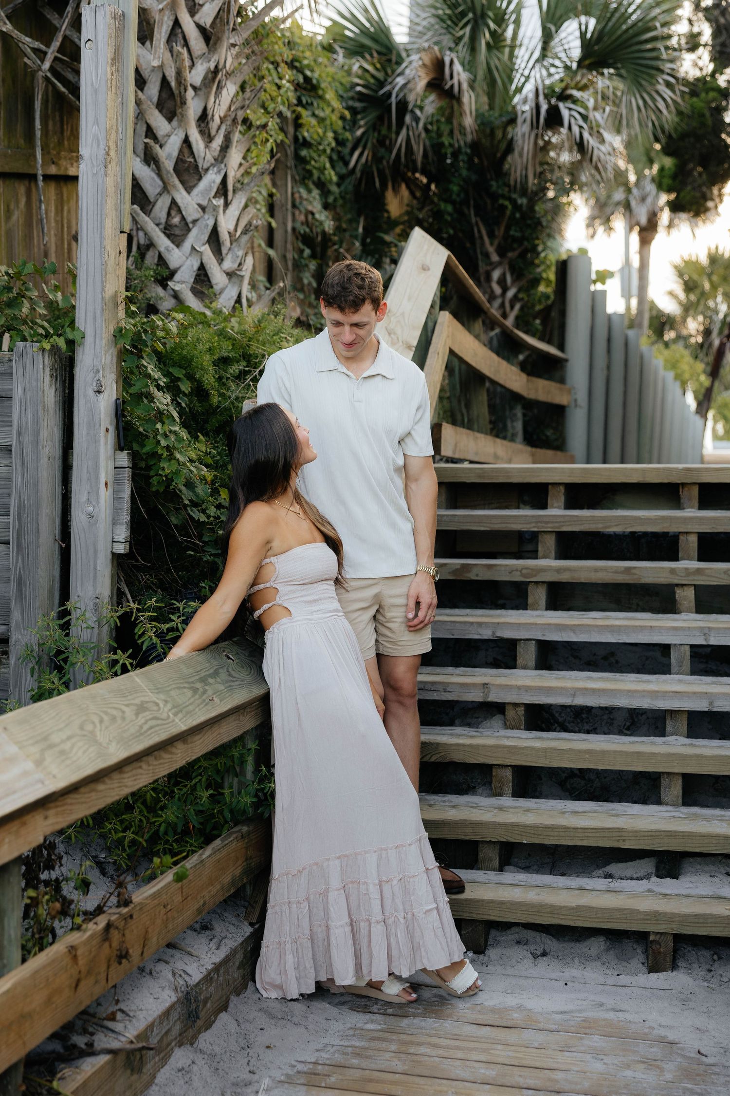 A couple in light colored outfits shares intimate moments on a wooden deck platform near palm trees.