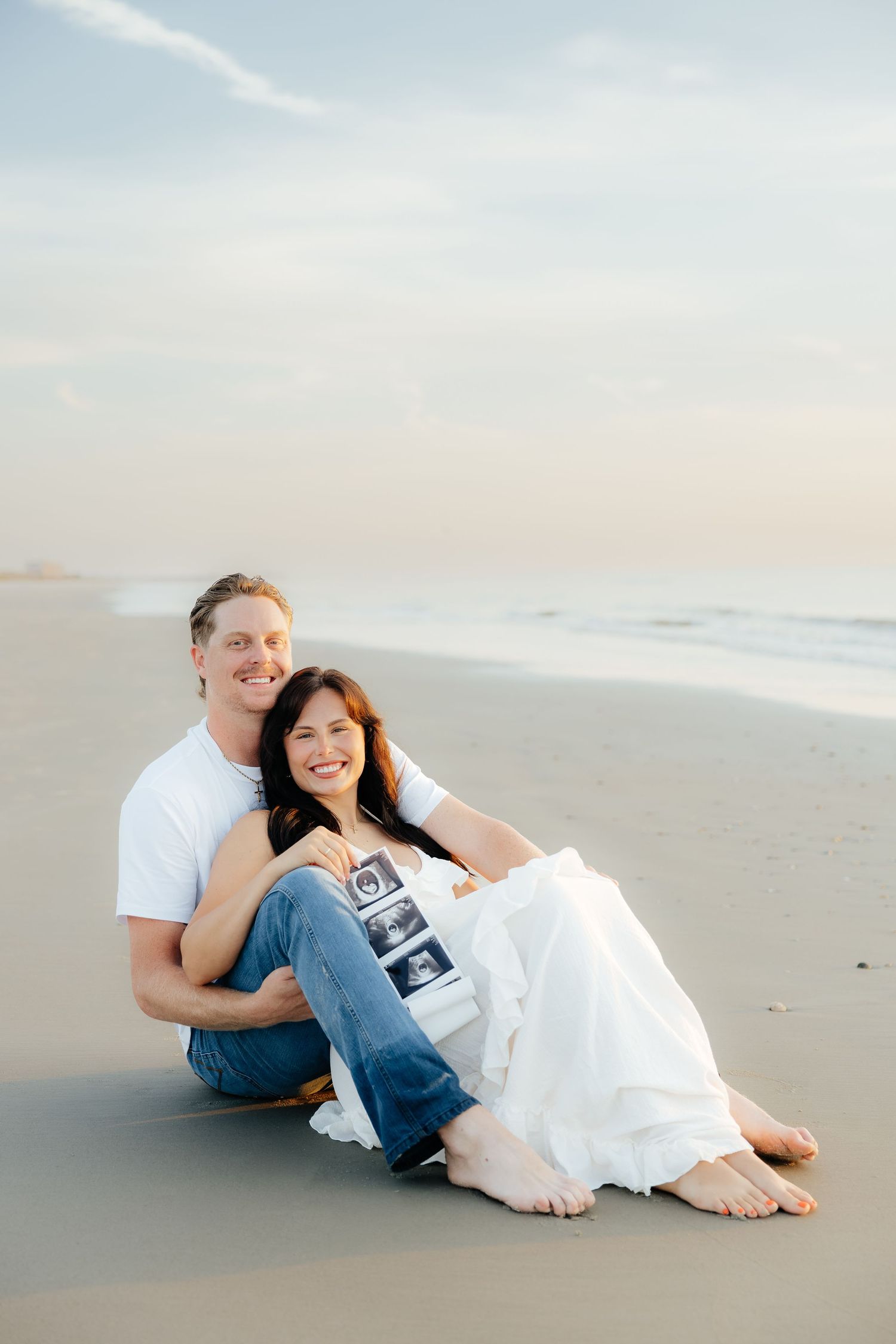 Happy couple sitting on beach holding ultrasound photos, woman in white dress, man in white shirt and jeans, ocean backdrop.