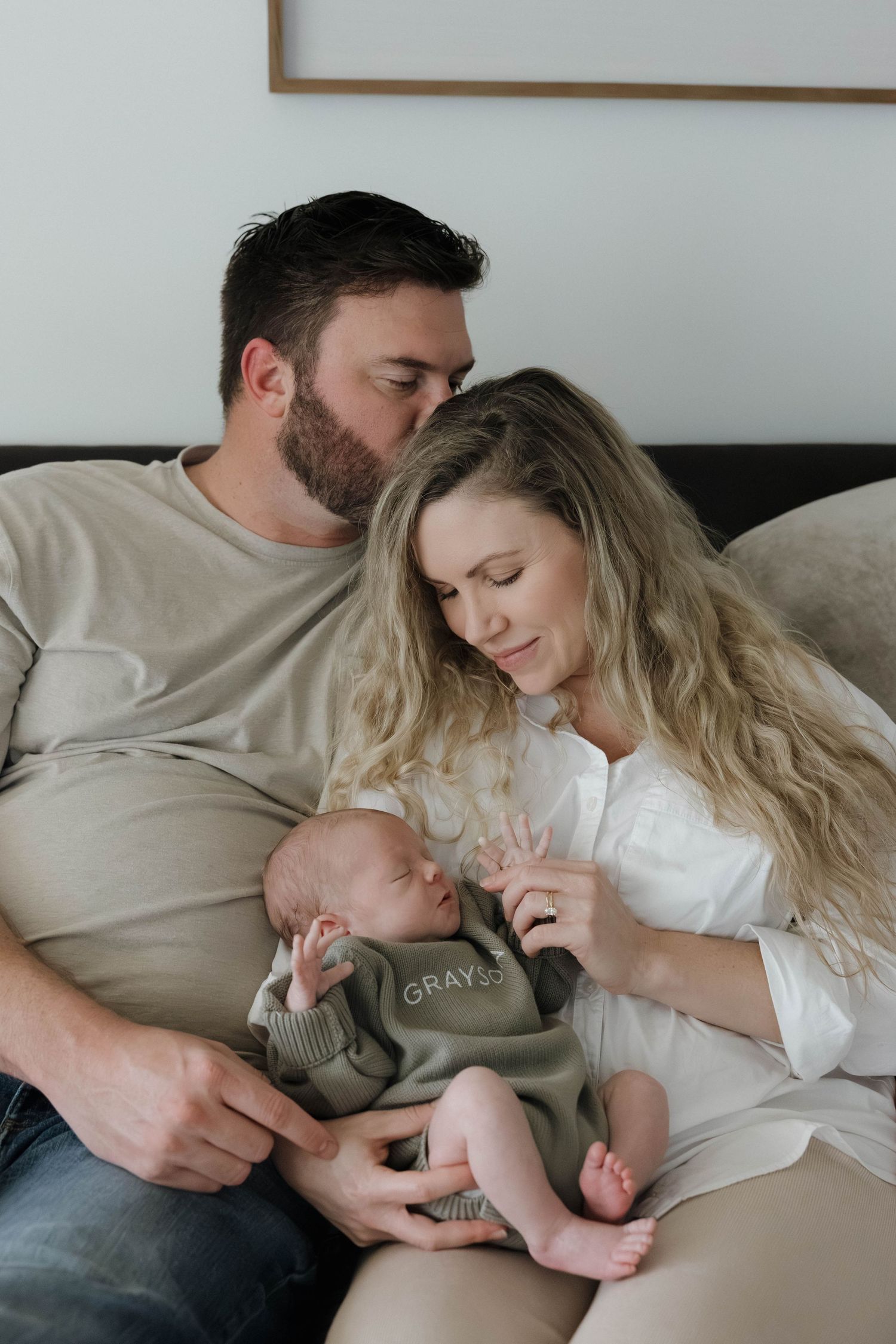 Tender family moment showing parents and baby resting together on white bedding in soft natural light.