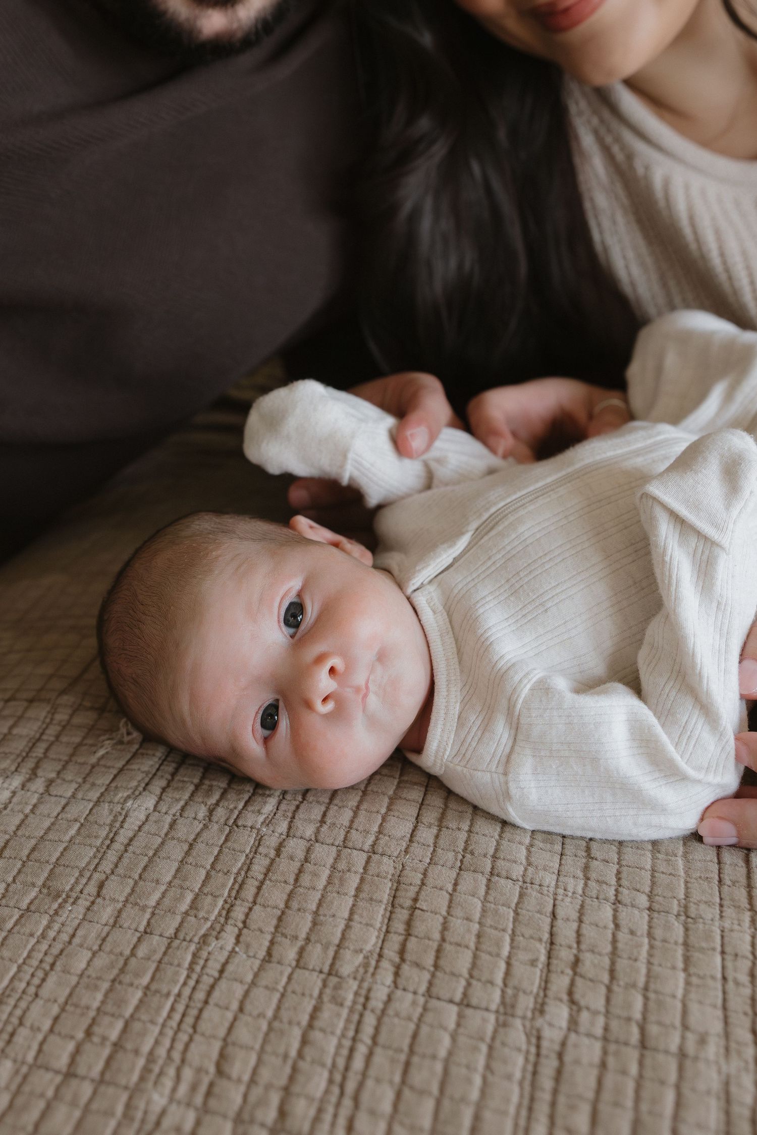 A baby wearing a white knit sweater lies on a textured beige surface looking at the camera.